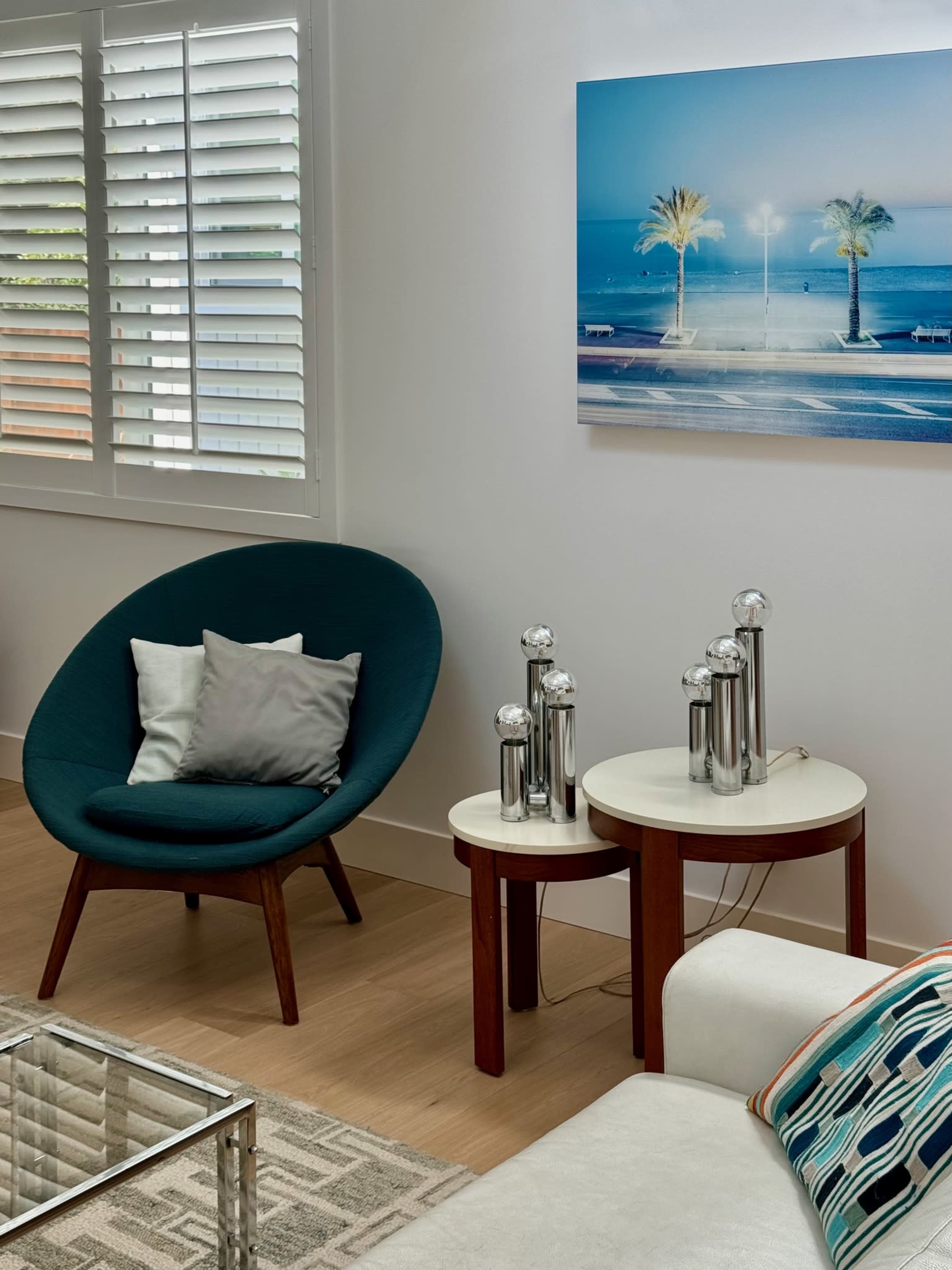 The image shows a modern living room with a green chair, two round side tables with metallic lamps, and a coastal-themed painting on the wall.