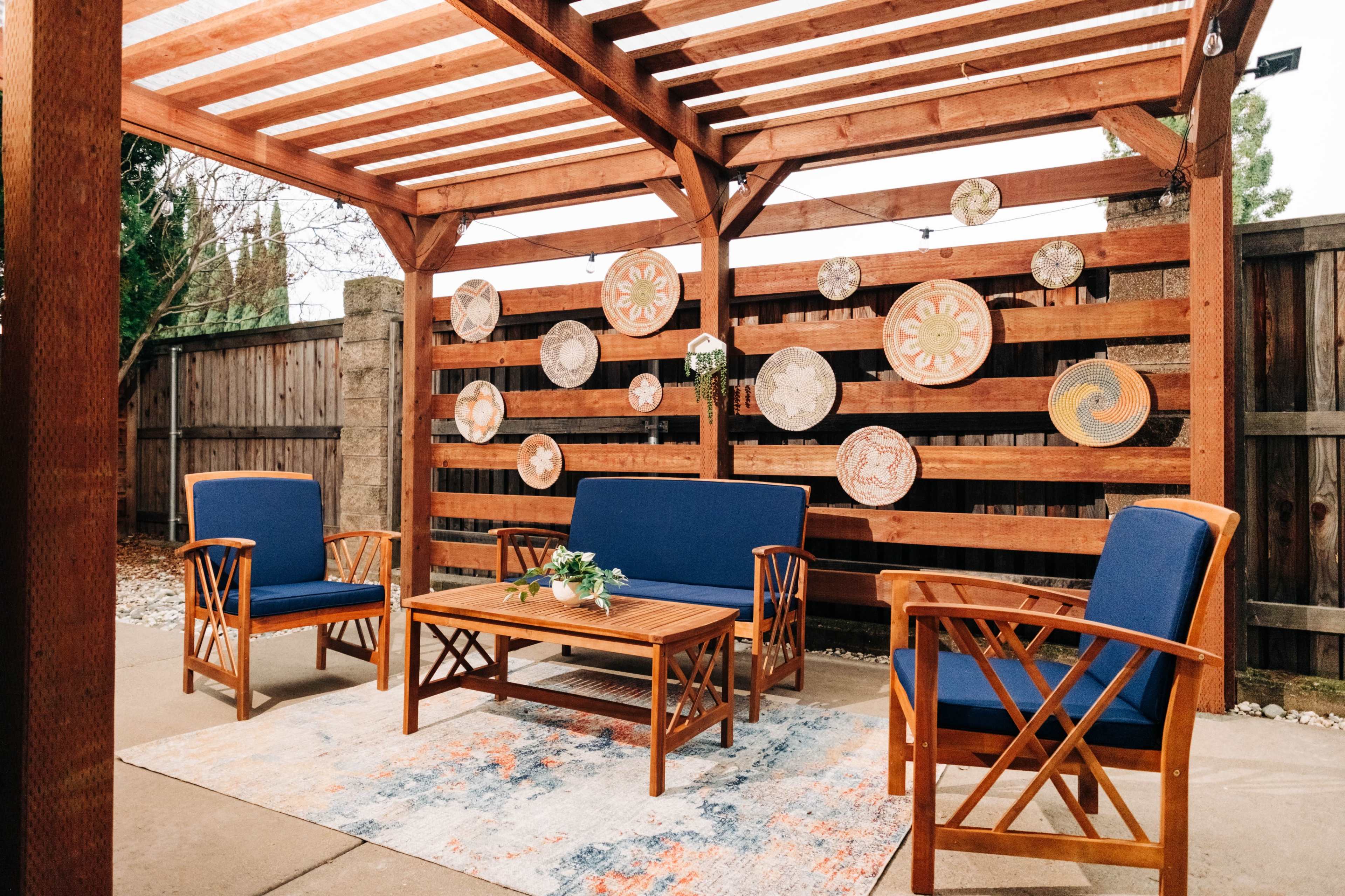 A wooden pergola with a slatted roof shelters a seating area featuring two blue chairs and a table, surrounded by decorative plates mounted on a wooden fence.