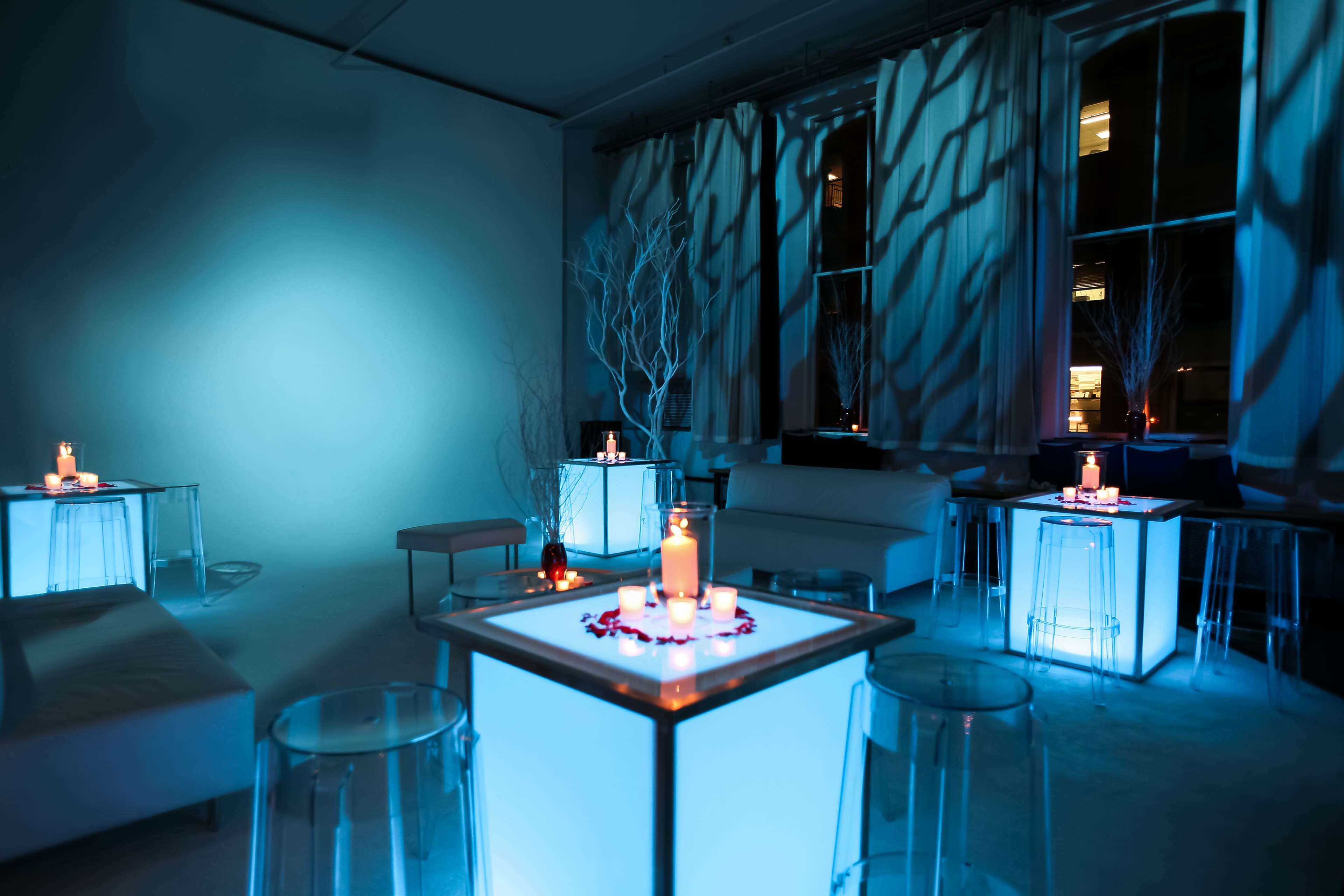 The scene features a dimly lit room with glowing blue tables adorned with candles and decorative elements, complemented by light filtering through sheer curtains.