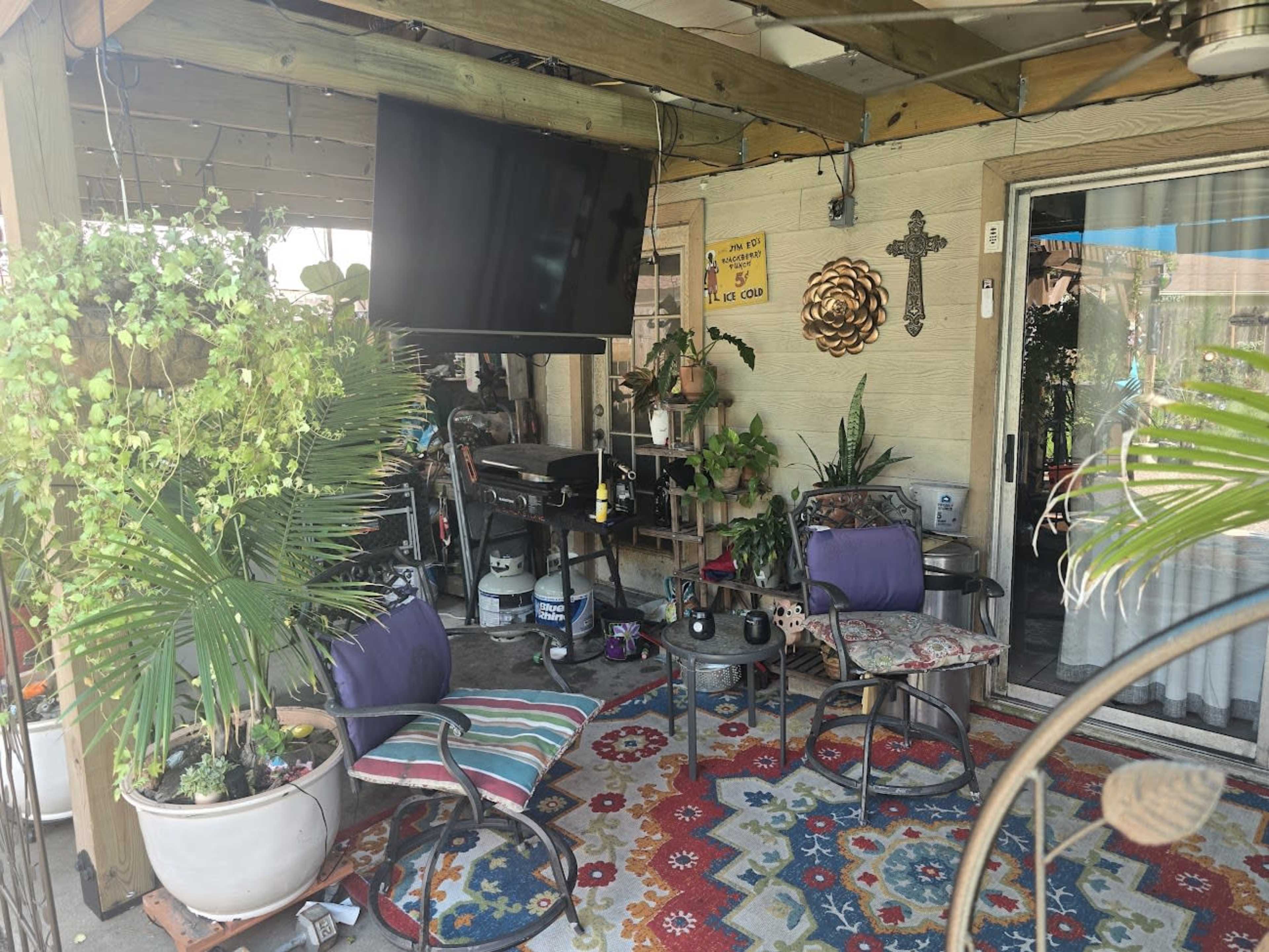 The image shows a cozy outdoor patio area with chairs, a colorful rug, potted plants, and a mounted television.