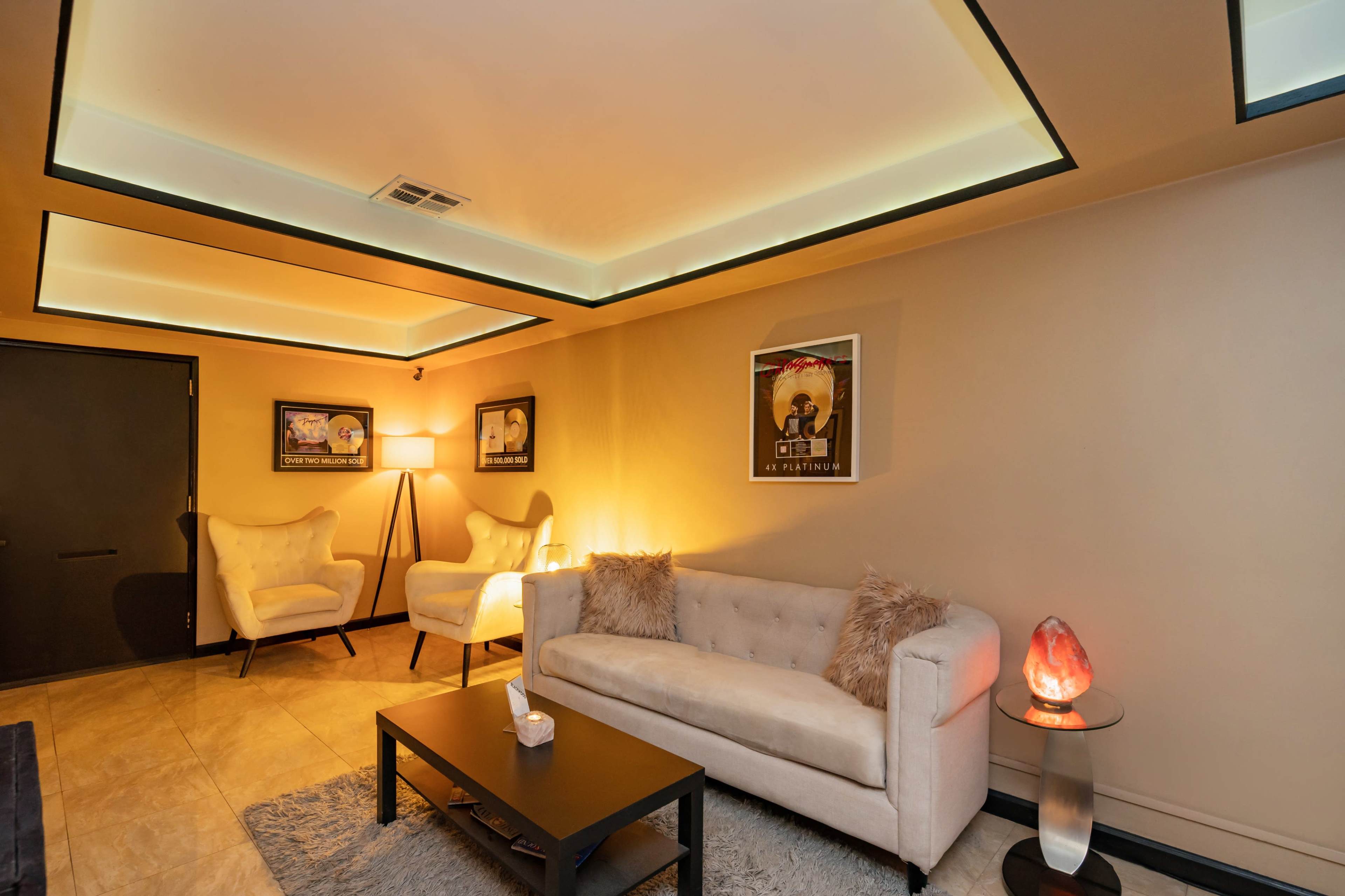 A cozy room with a light-colored sofa, two armchairs, a coffee table, and framed artwork on the walls, illuminated by soft lighting.