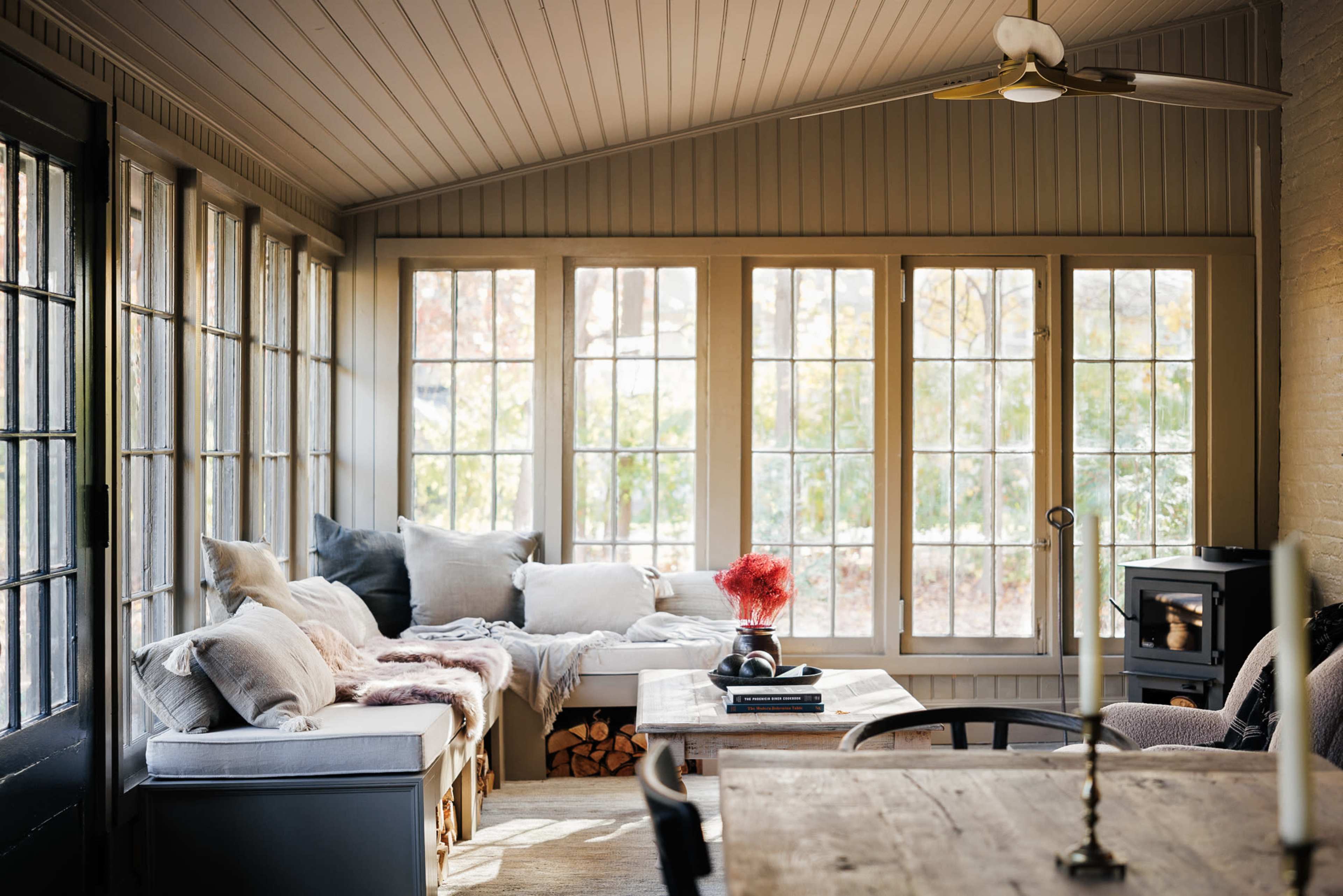 A sunlit room with large windows, a wood stove, a table, and cozy seating areas filled with pillows and blankets.
