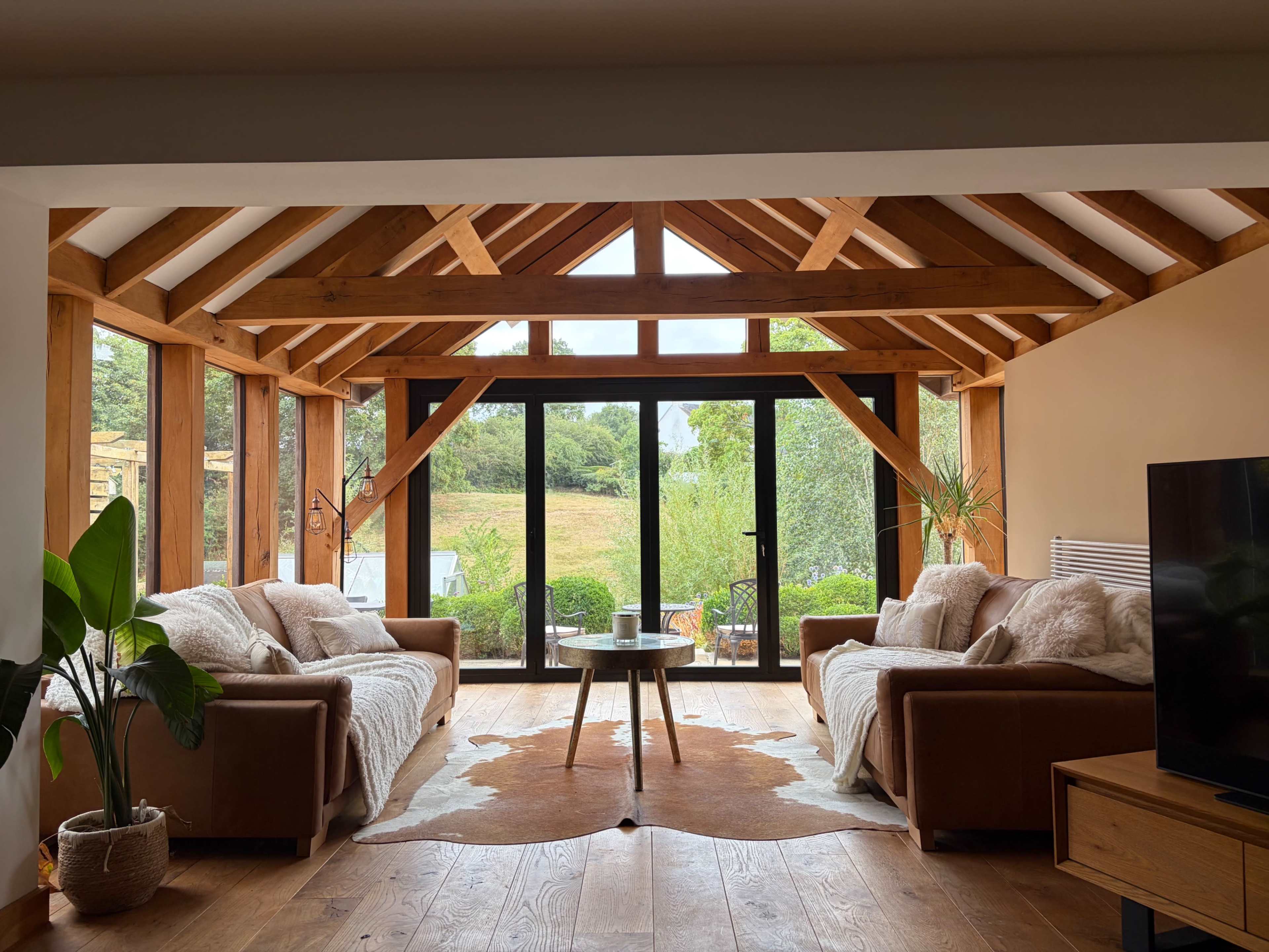 The living room features two brown sofas facing a coffee table, with large windows framed by wooden beams that provide a view of the outdoor landscape.