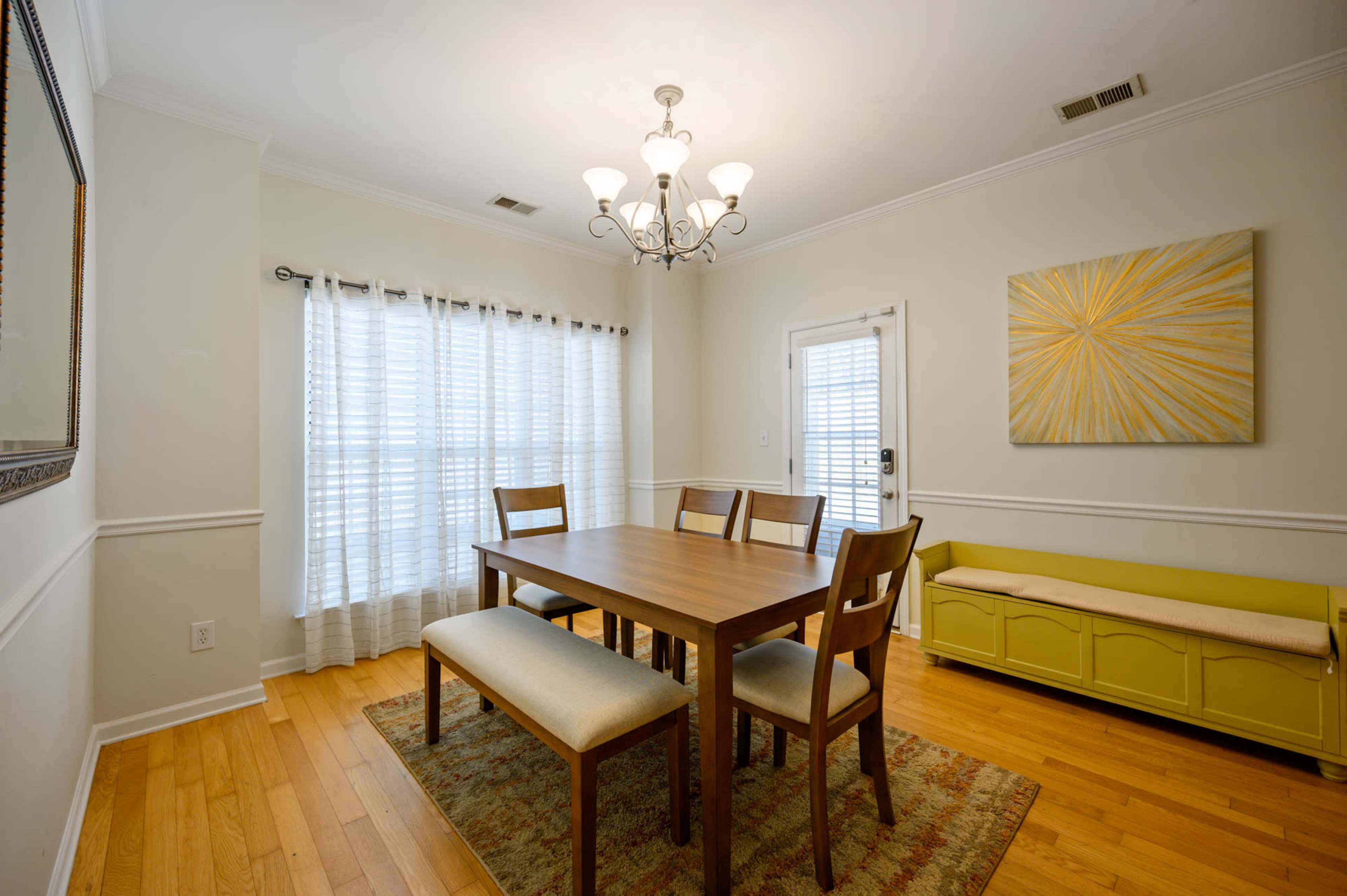 A dining room features a wooden table surrounded by four chairs, with a chandelier above and a bench against a wall, illuminated by natural light through sheer curtains.