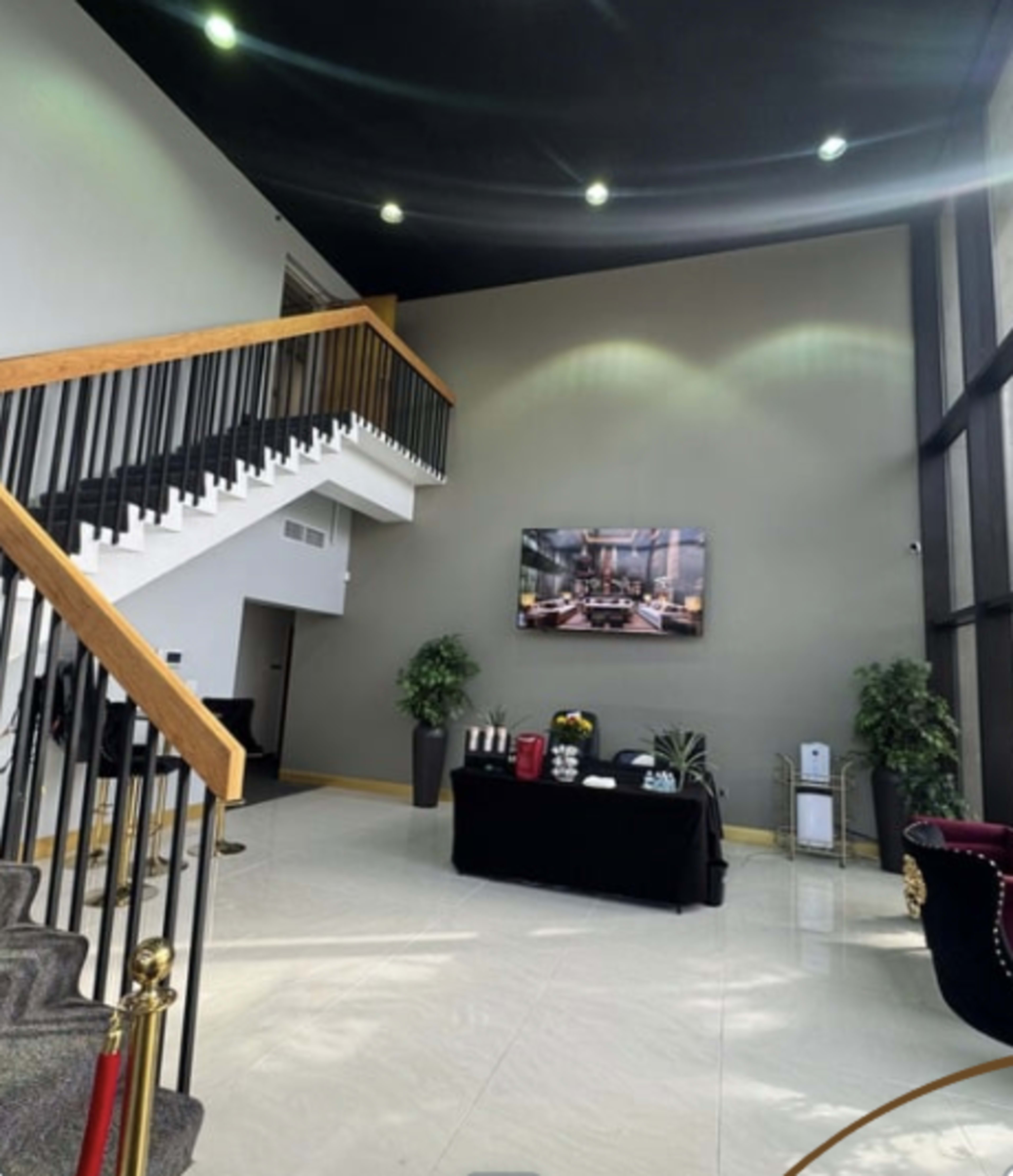 The image shows a spacious lobby with a staircase, a television mounted on the wall, and plants in decorative pots.
