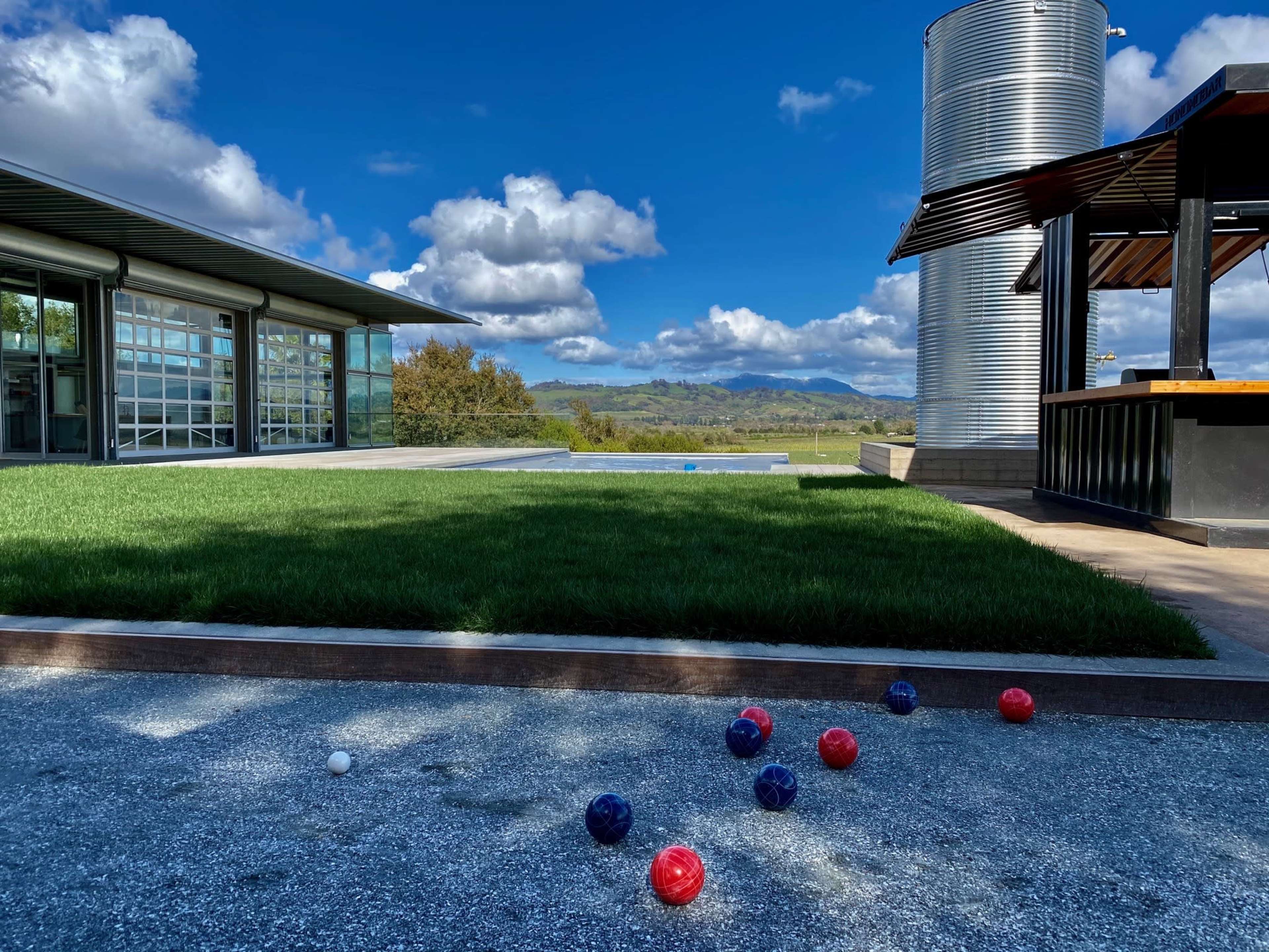 A landscaped area with a lawn, a bocce ball court featuring scattered balls, and a modern building alongside a silver water tank, set against a backdrop of mountains and blue sky.