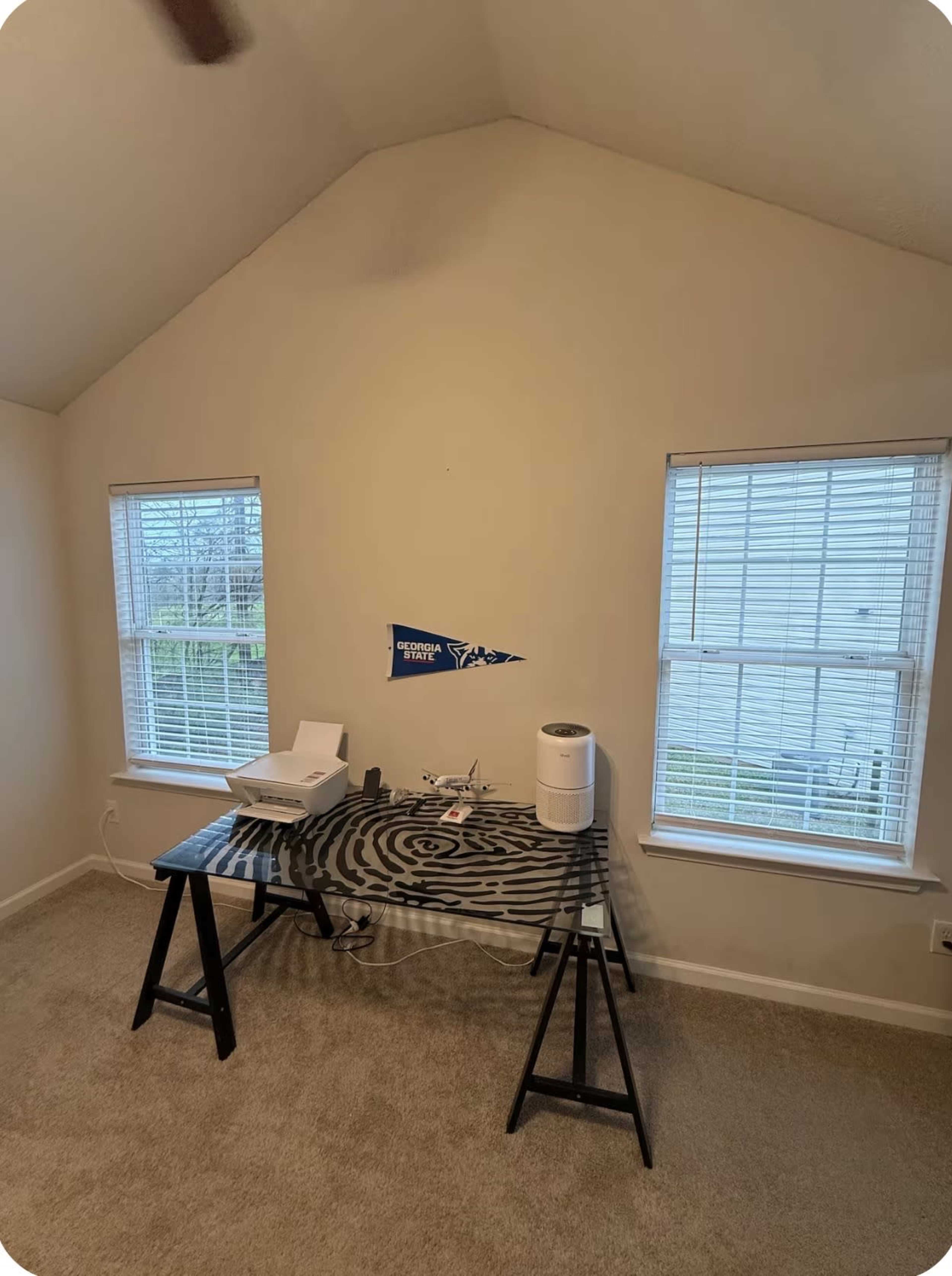 The image shows a room with a slanted ceiling, featuring a desk, a printer, and two windows that allow natural light to enter.