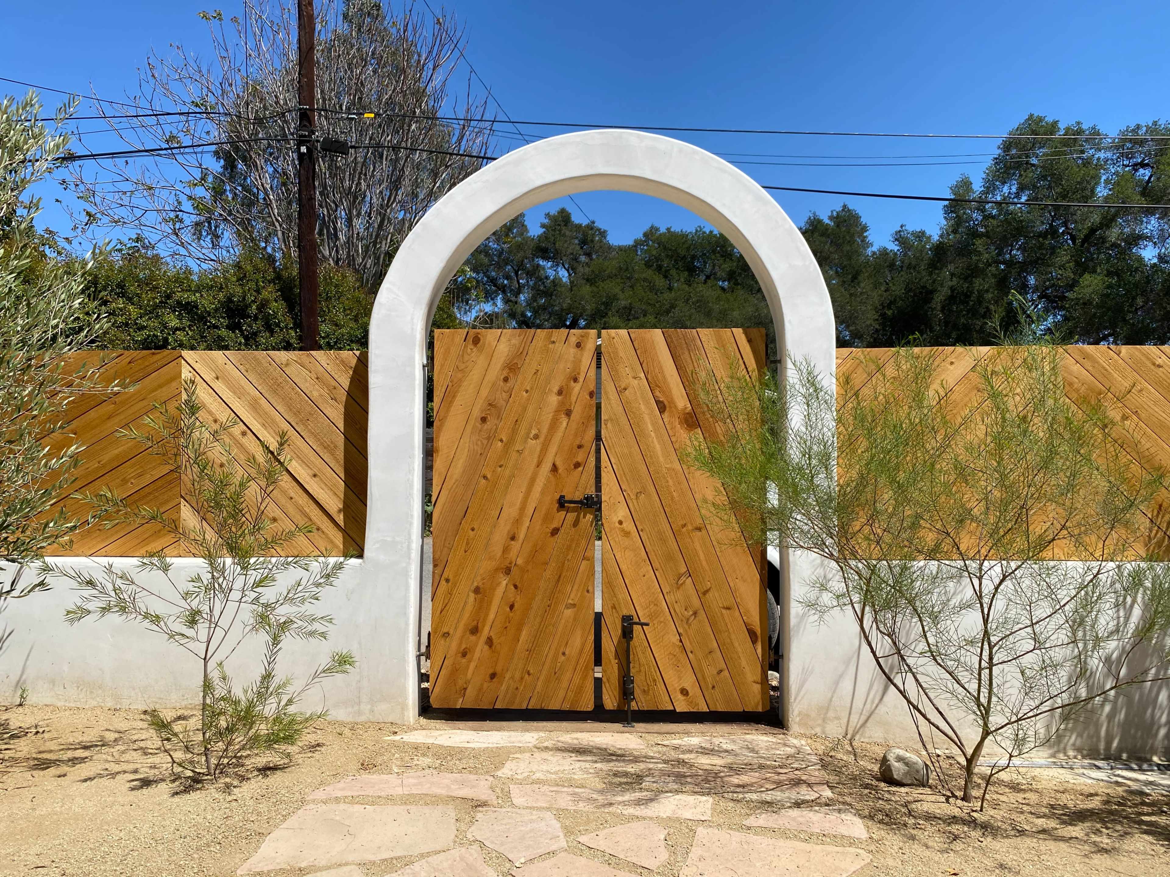A wooden gate with an archway is set within a white stucco wall, surrounded by landscaped gravel and plants.