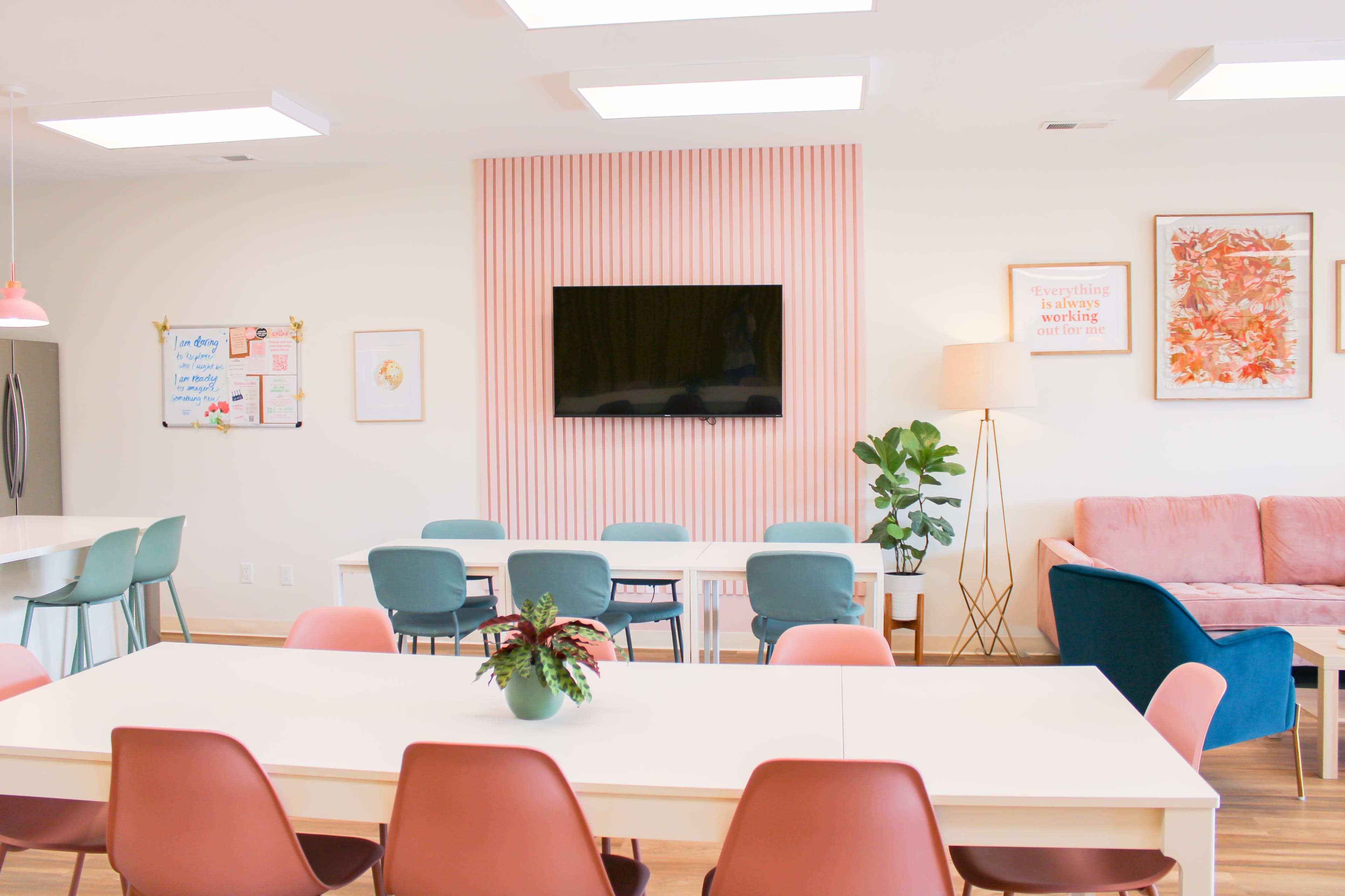 A bright, modern communal space features tables, chairs, a television on a pink accent wall, and a cozy seating area with a sofa and plants.