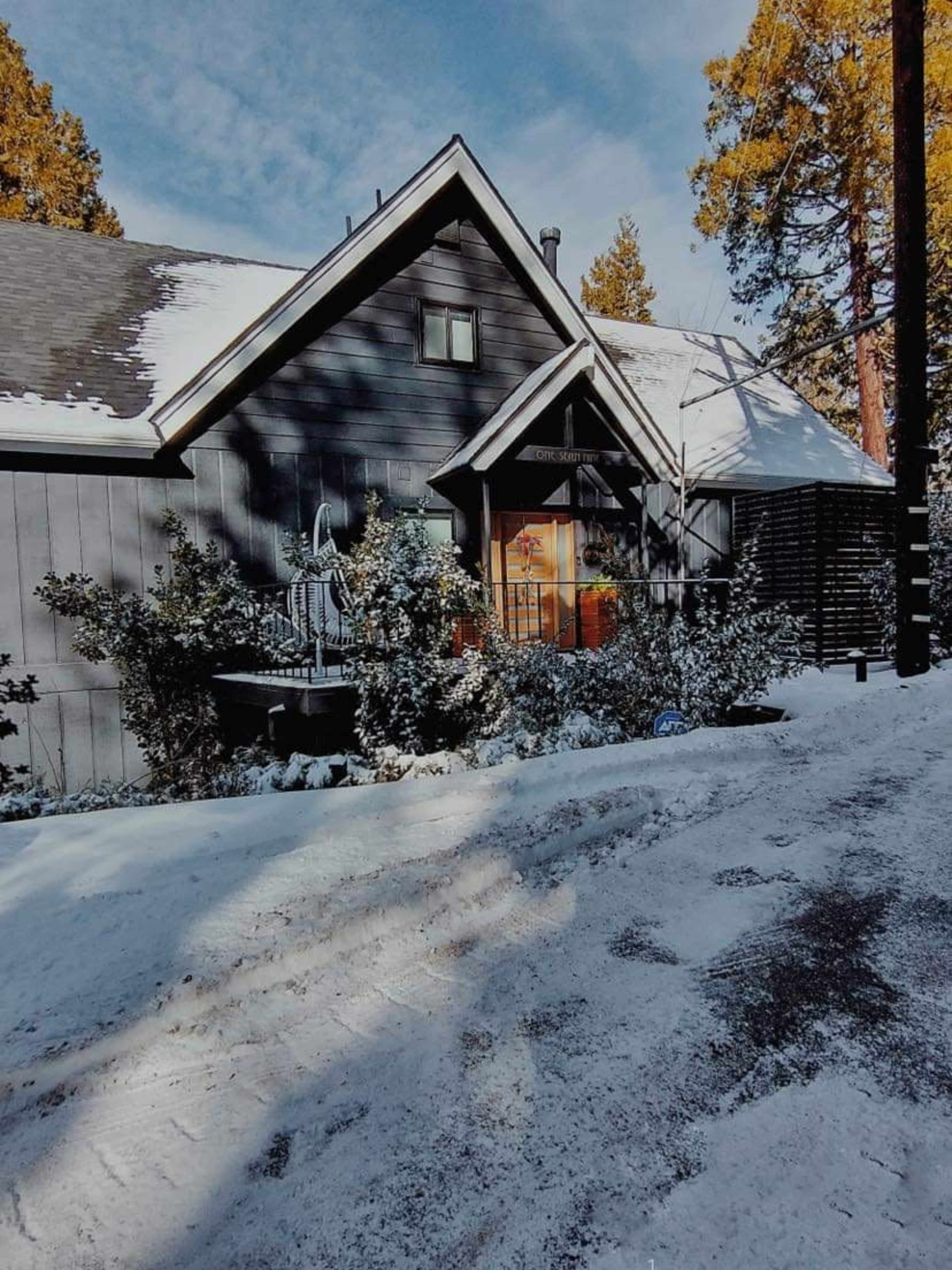 A dark wooden house is surrounded by snow-covered ground and trees, with a clear blue sky overhead.