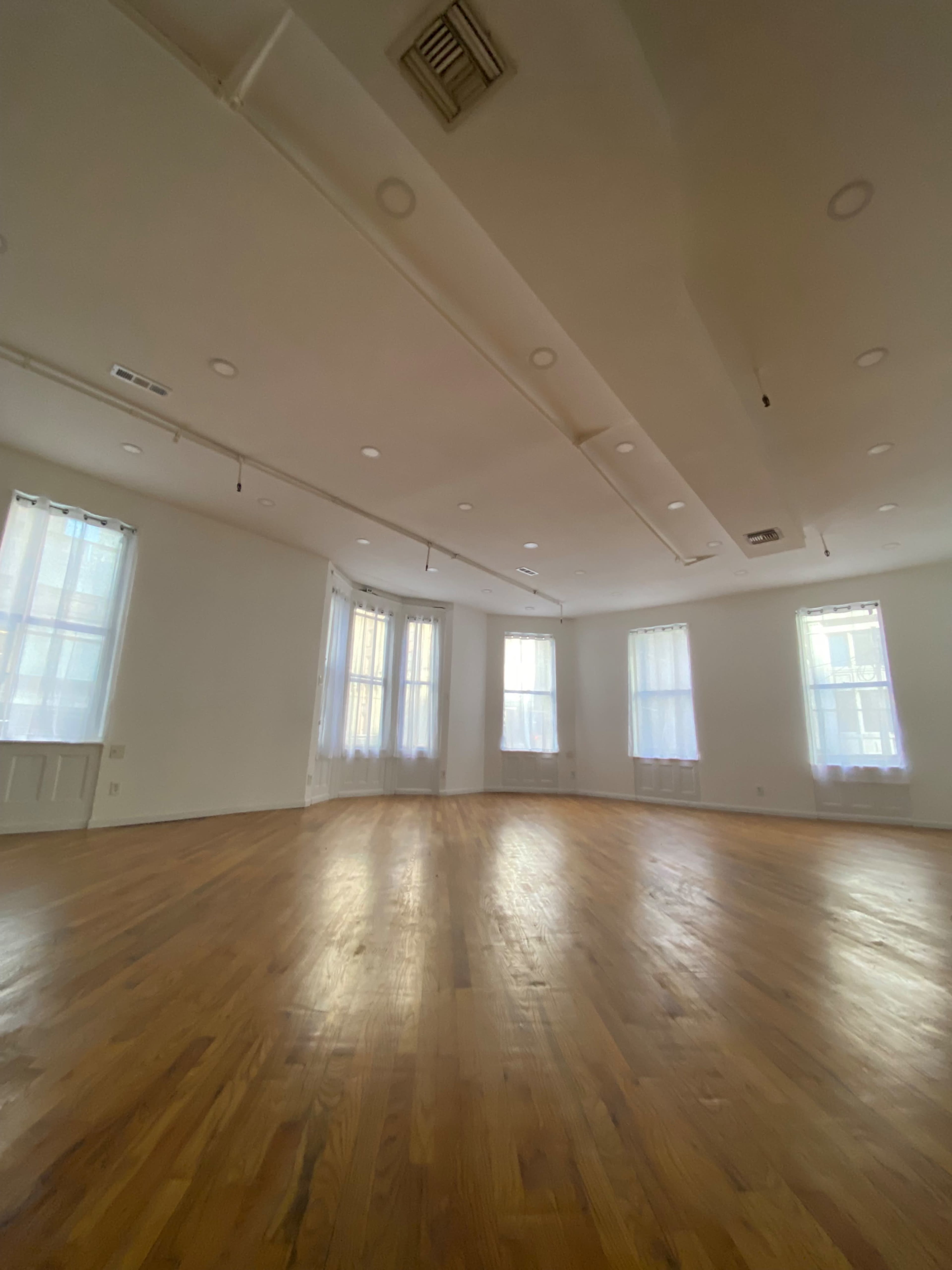 The image shows a spacious room with hardwood floors, large windows, and clear walls.