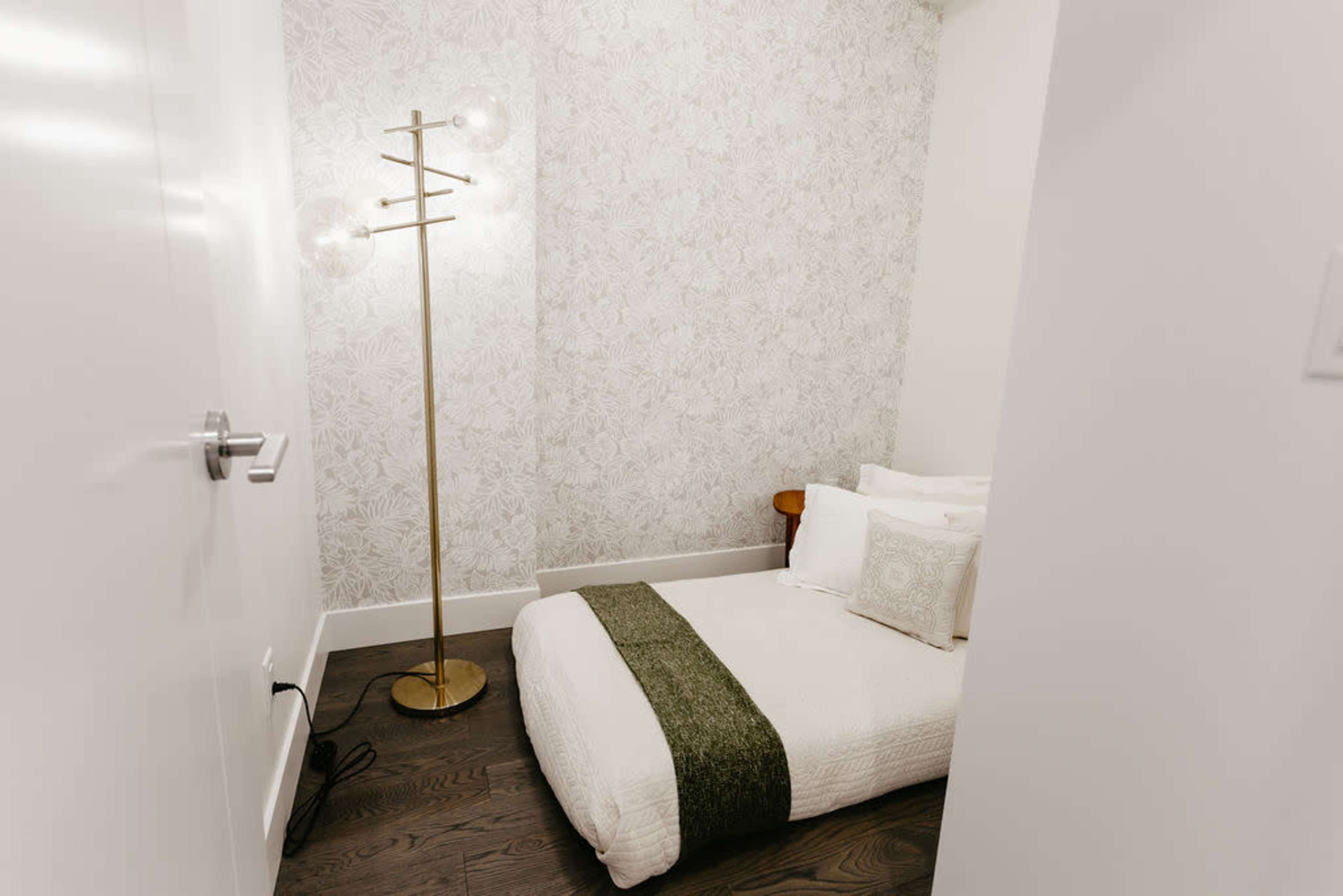 A small room features a bed with a green throw, a floor lamp, and floral-patterned wallpaper.