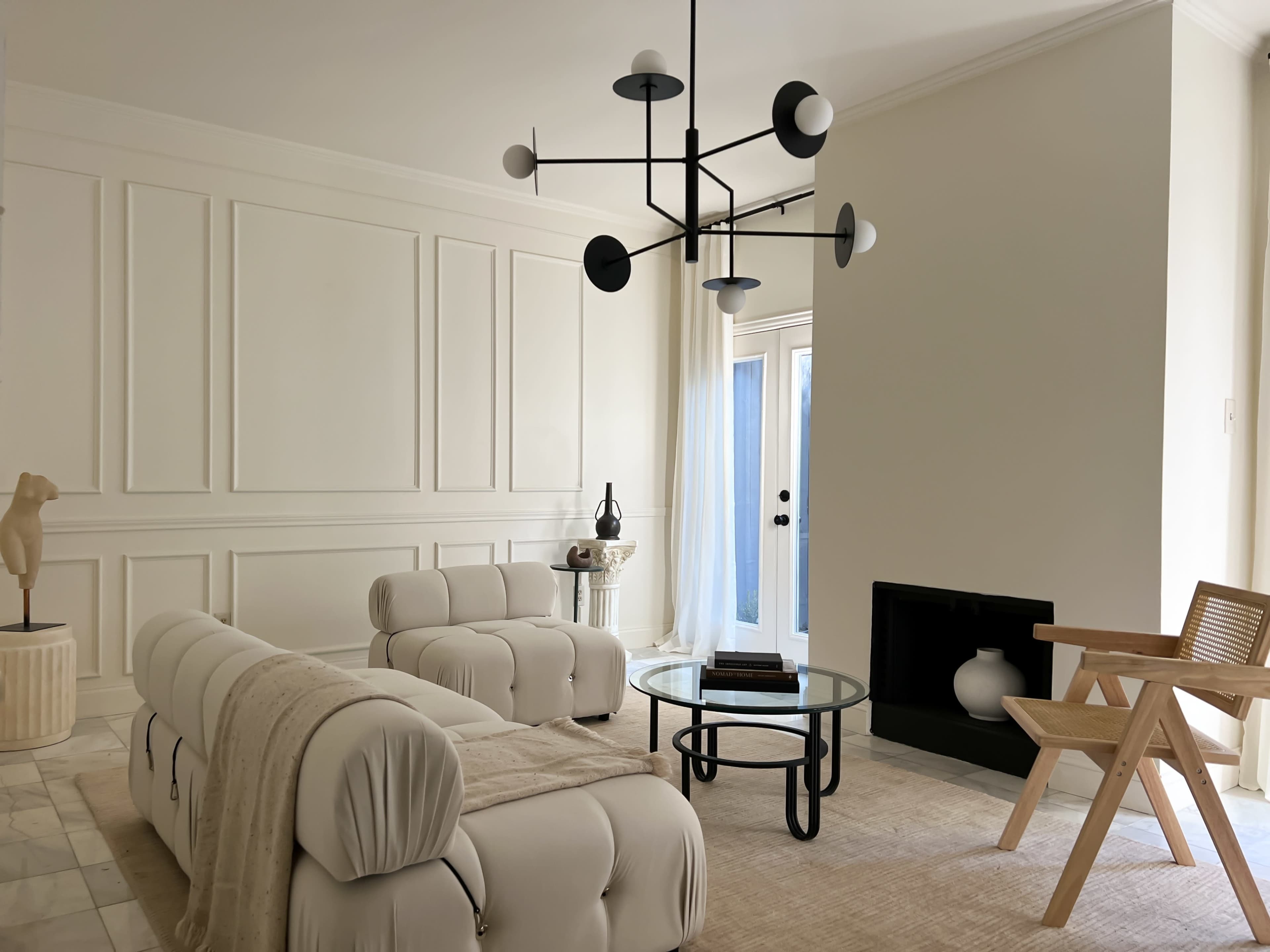 A minimalist living room features a pair of white sofas, a glass coffee table, a wooden chair, and a modern chandelier against a softly lit backdrop with a sliding glass door.