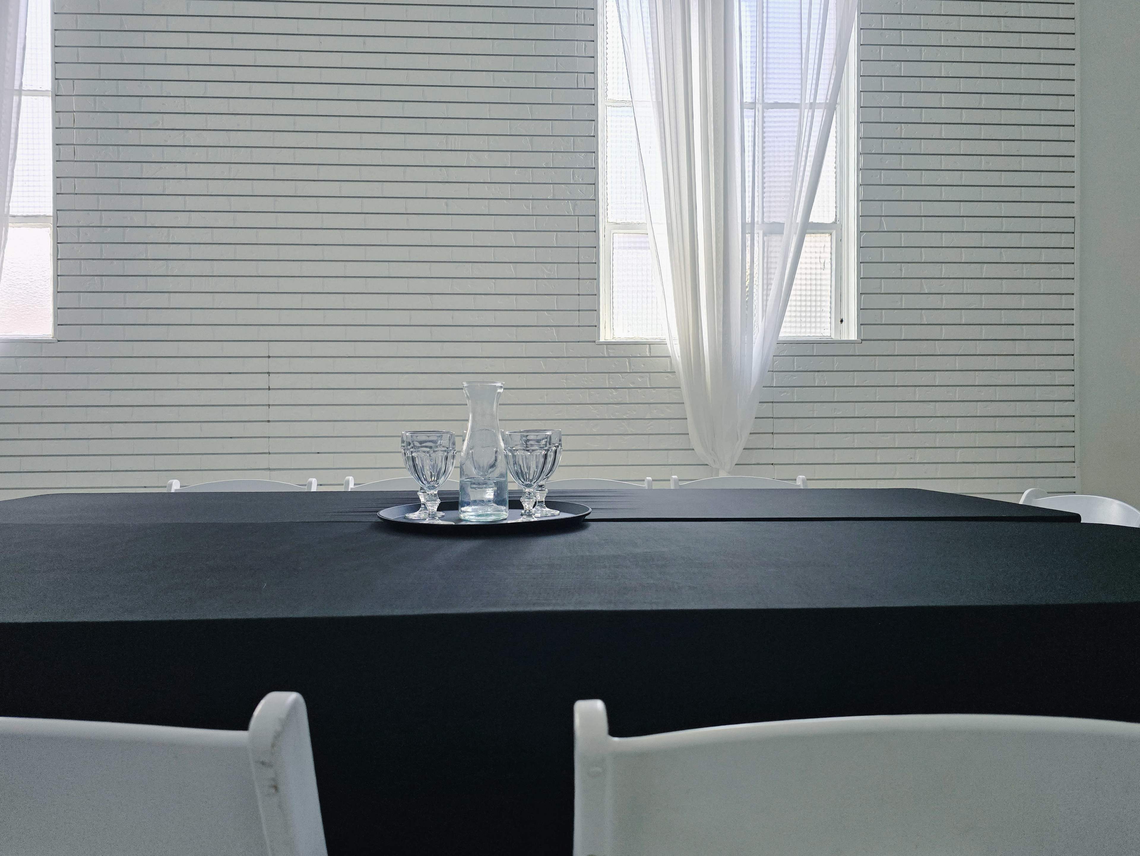 A black tablecloth covers a table set with a clear glass pitcher and three glasses on a tray, with white curtains and a light-colored wall in the background.