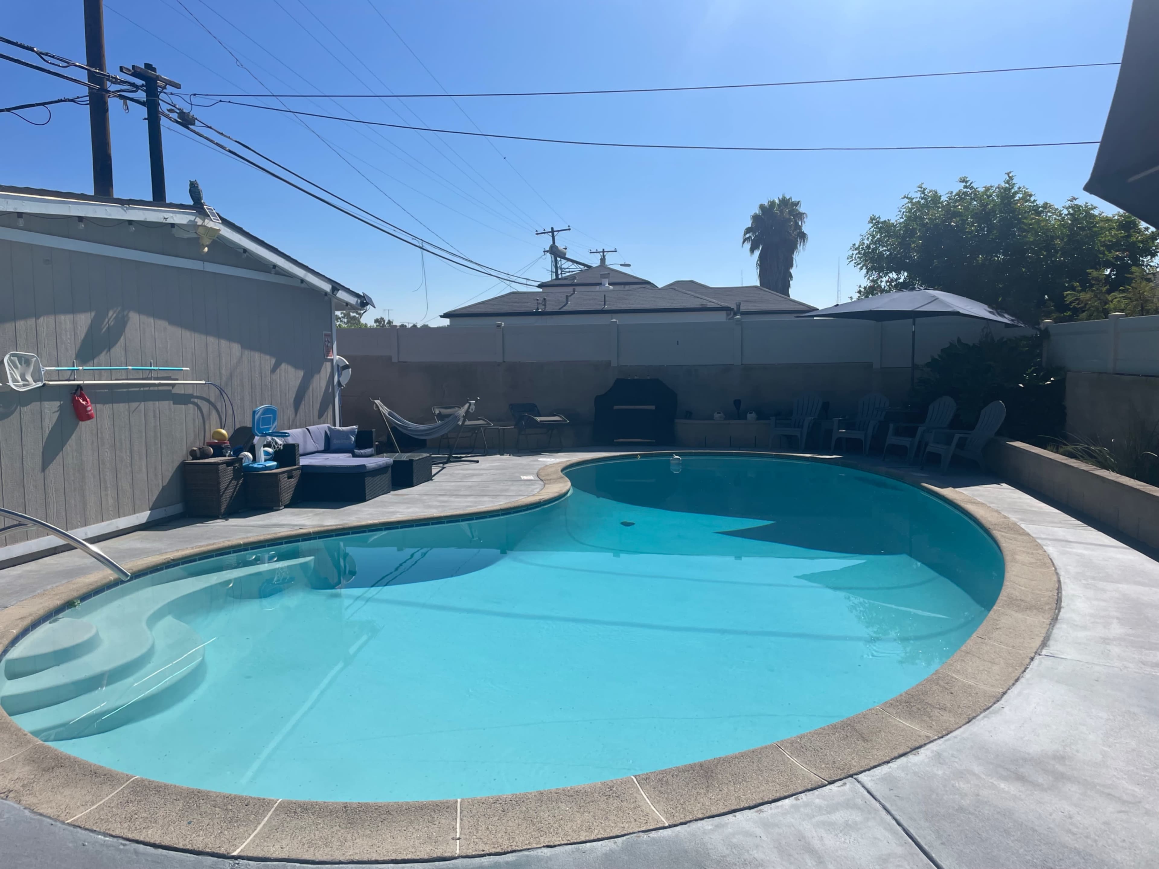 A swimming pool surrounded by a concrete patio, various outdoor seating arrangements, and residential buildings in the background under a clear blue sky.