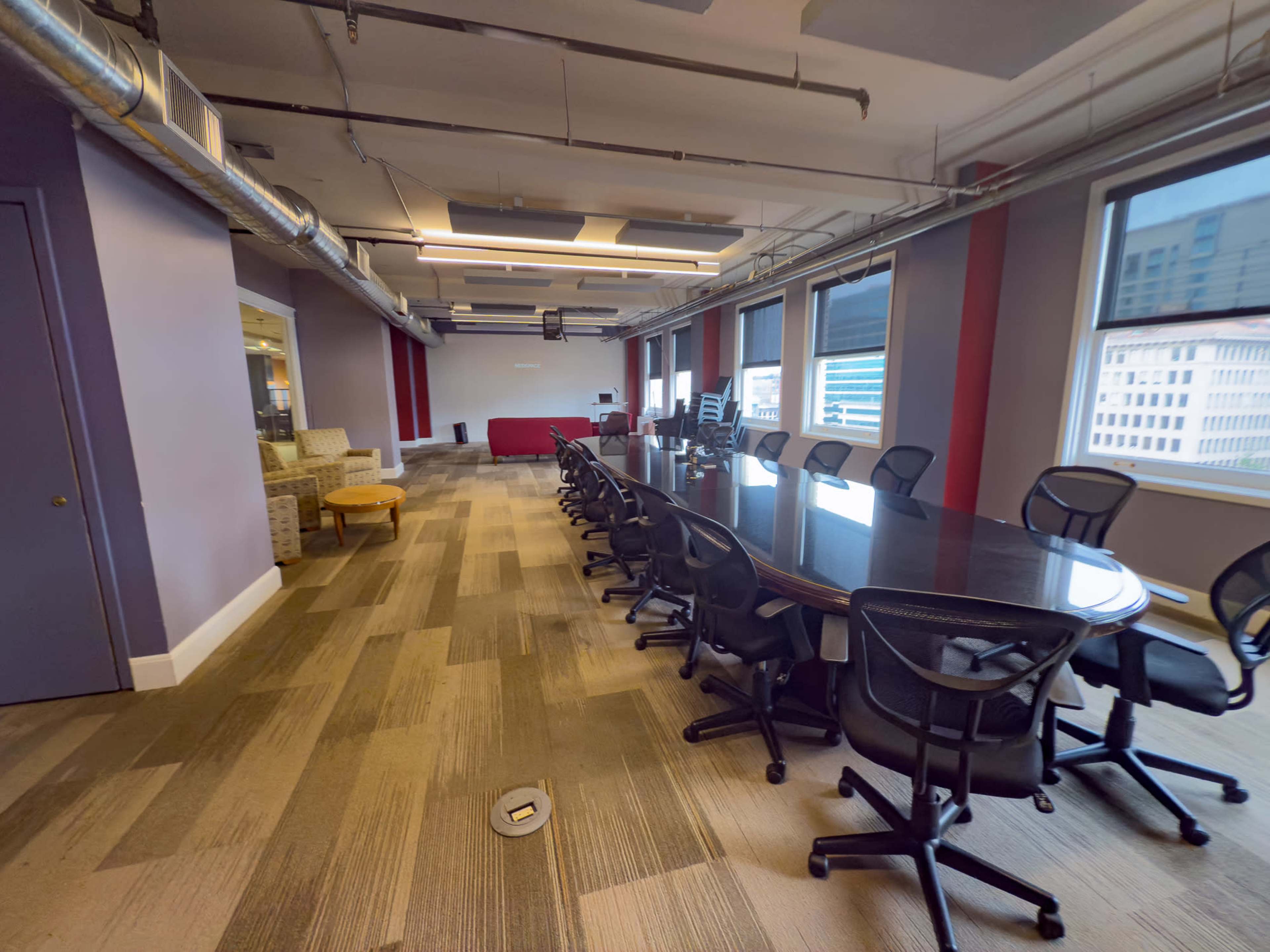 A large conference room features a long, dark wooden table surrounded by ergonomic chairs, with floor-to-ceiling windows providing natural light.