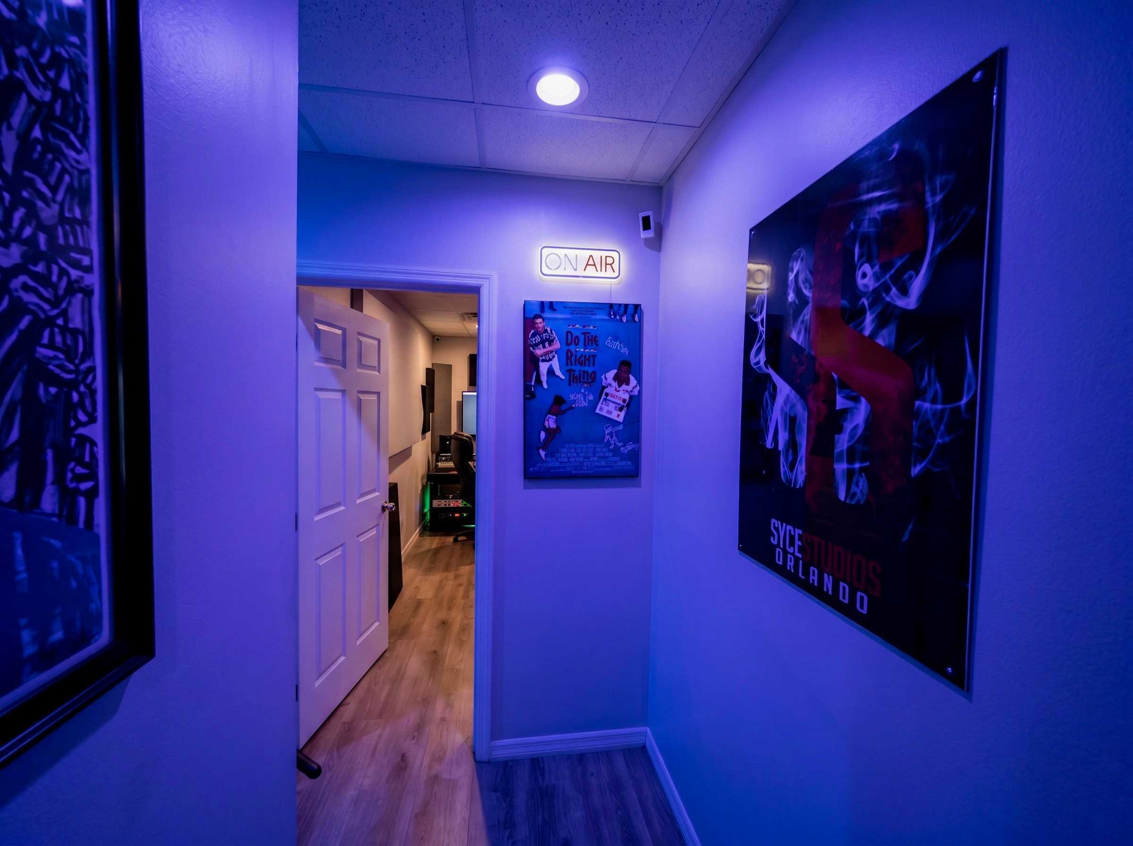 A dimly lit hallway features a neon "ON AIR" sign above a door, flanked by posters on the walls and leading to another room.