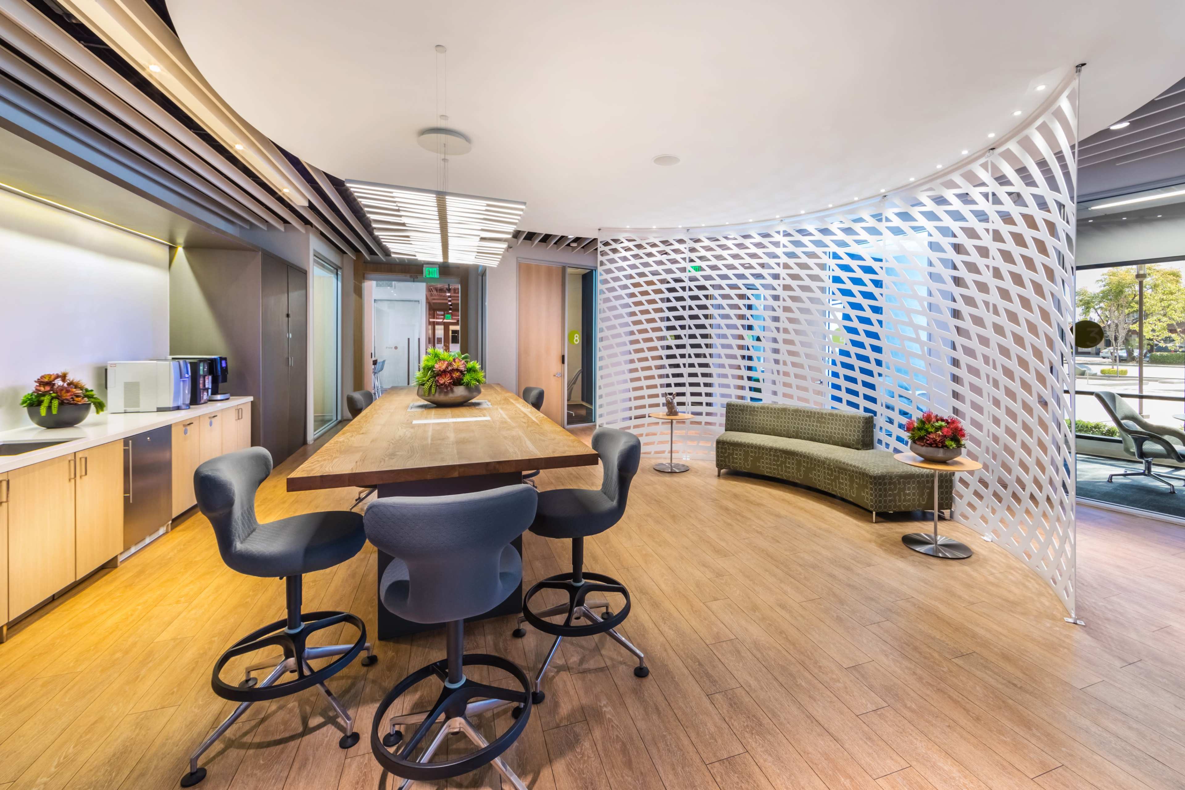 A modern office space with a wooden table surrounded by high stools, a curved seating area, and decorative plants.