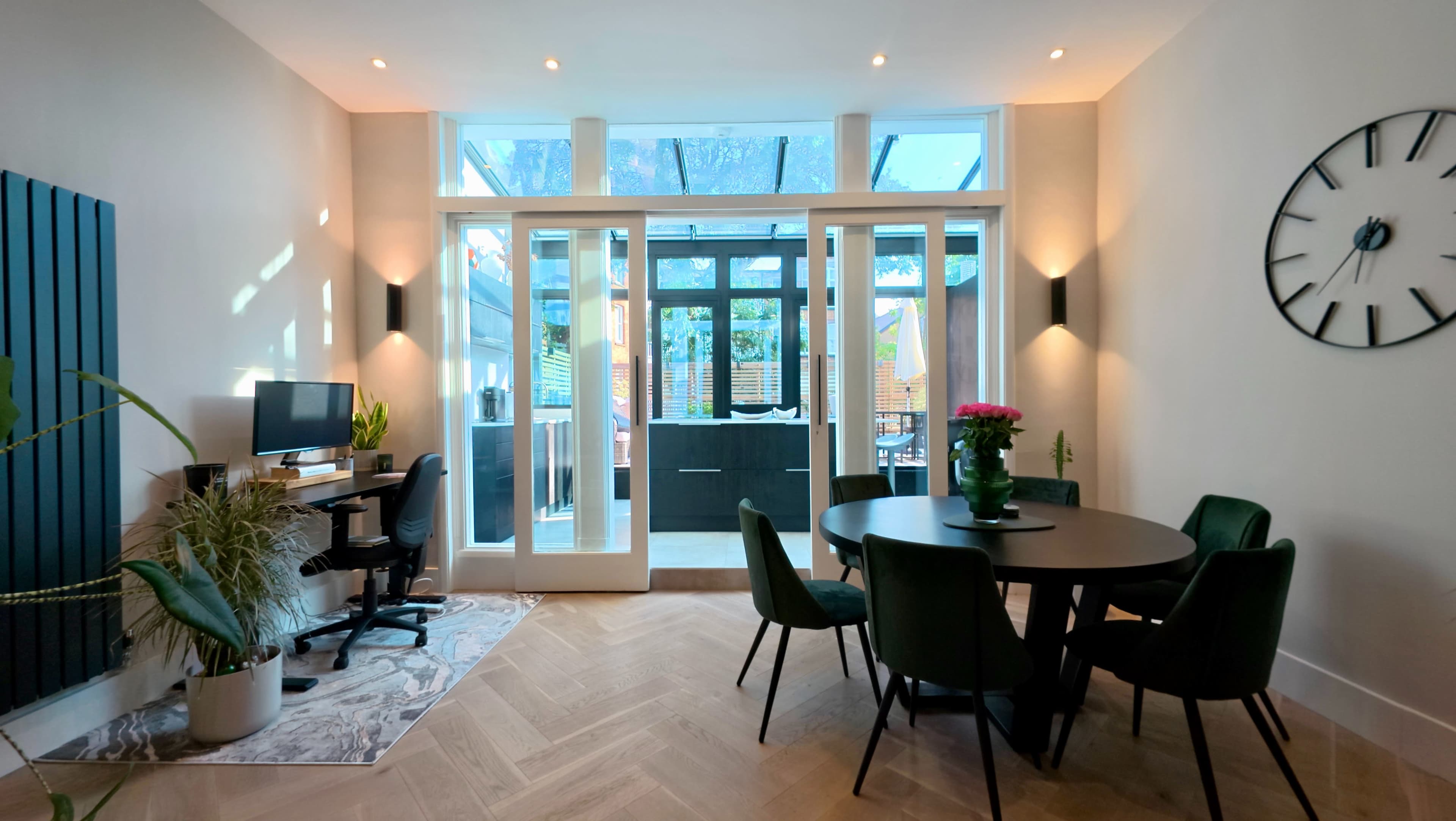 A modern interior features a dining area with a round table and chairs, a small workspace with a computer, and large windows allowing natural light to enter.