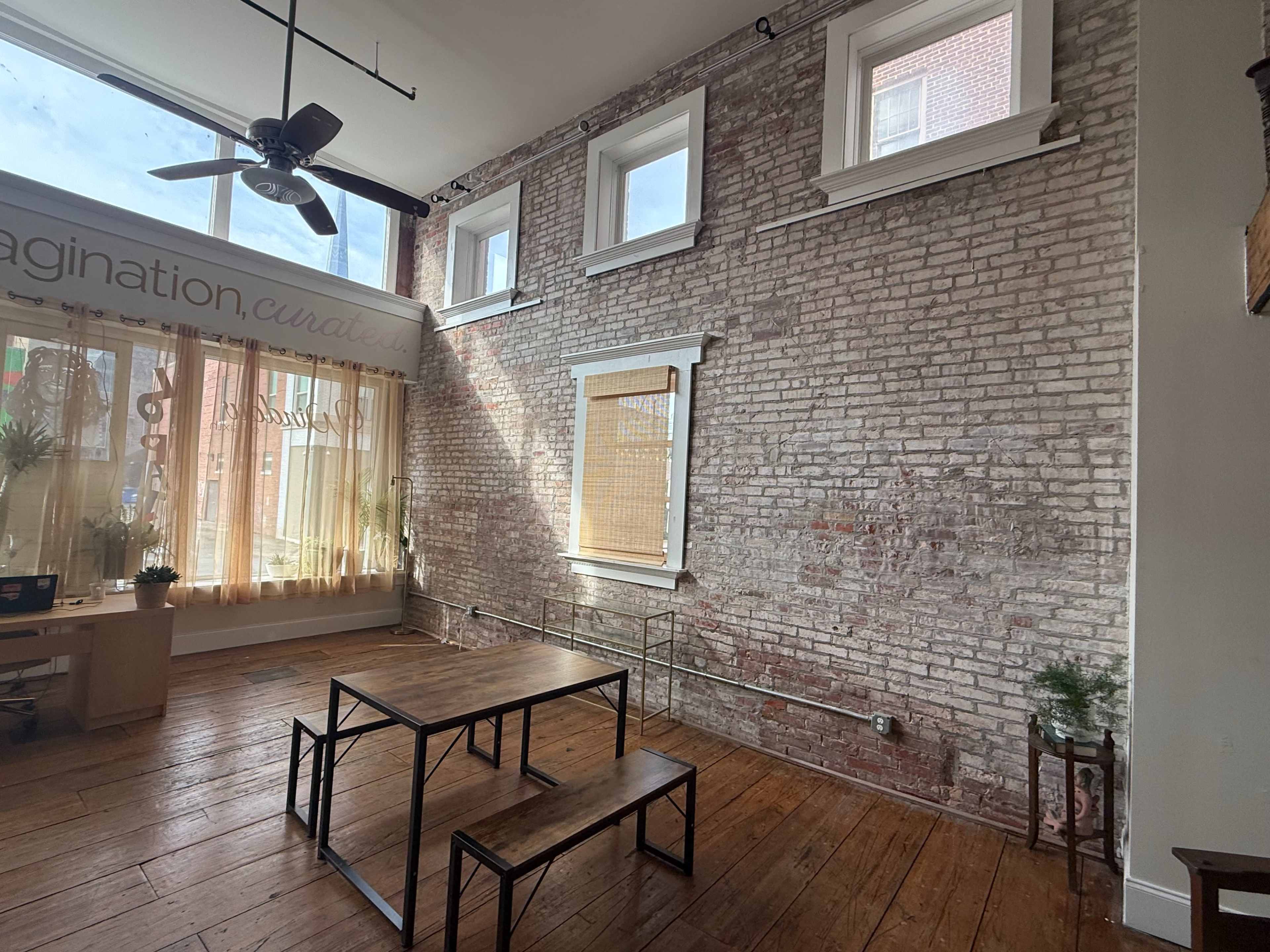 The room features exposed brick walls, large windows, and a simple wooden table set on a wooden floor.