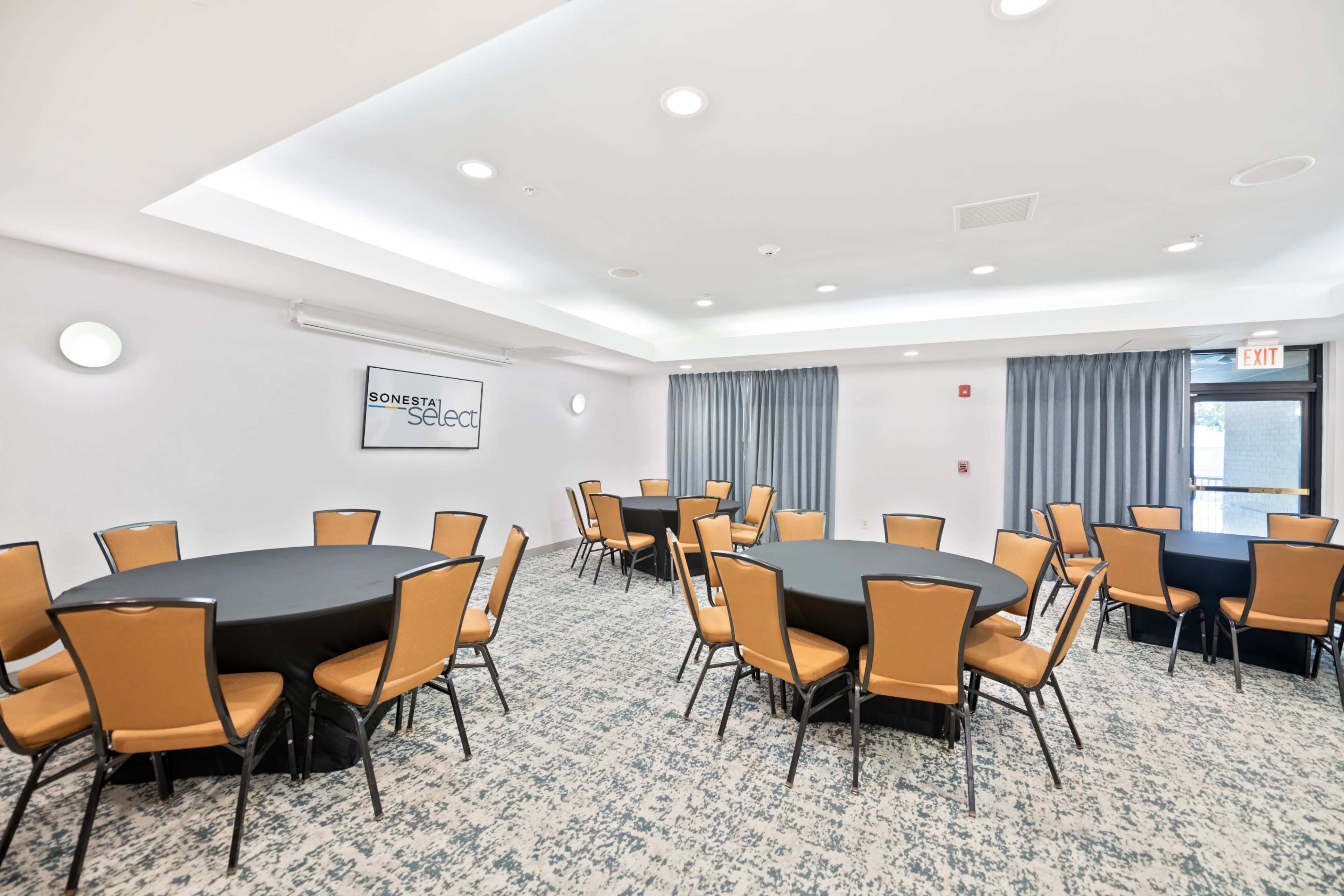 The image shows a meeting room with multiple round tables covered in black cloth and surrounded by orange chairs, featuring windows with gray curtains.