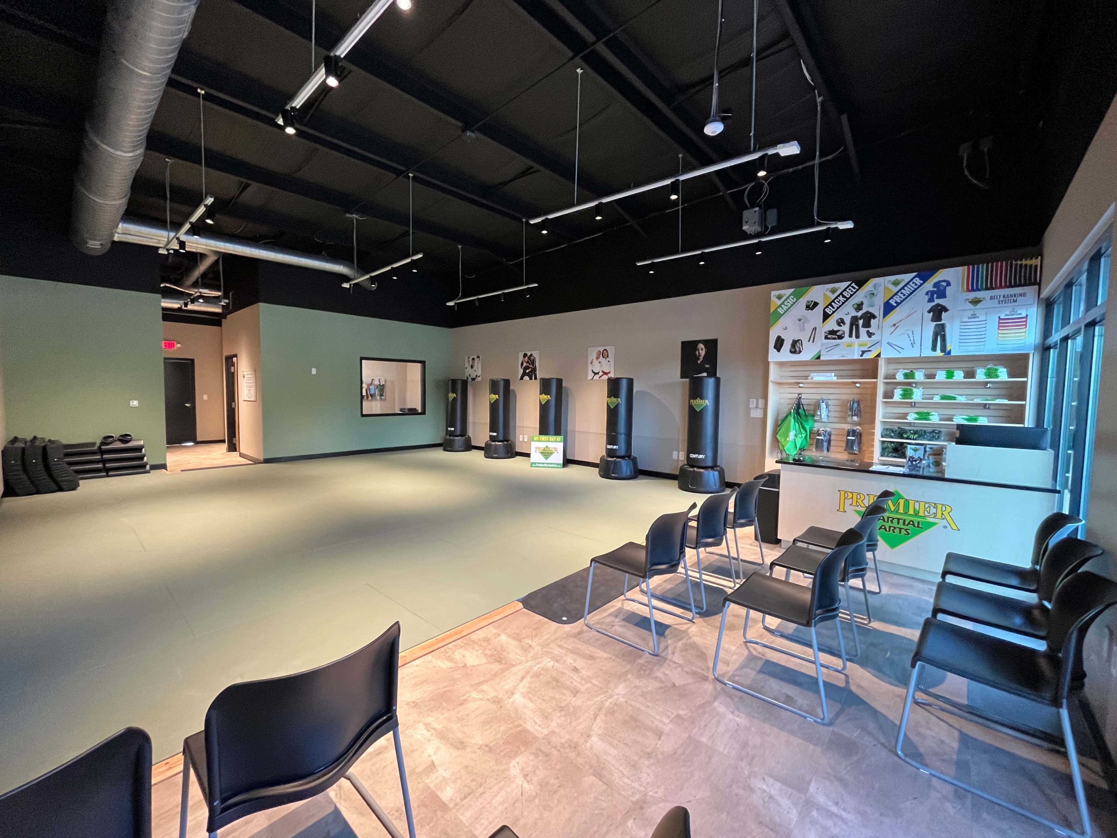 The image shows an empty martial arts studio with mats on the floor, a seating area with chairs, and various training equipment displayed on a wall.