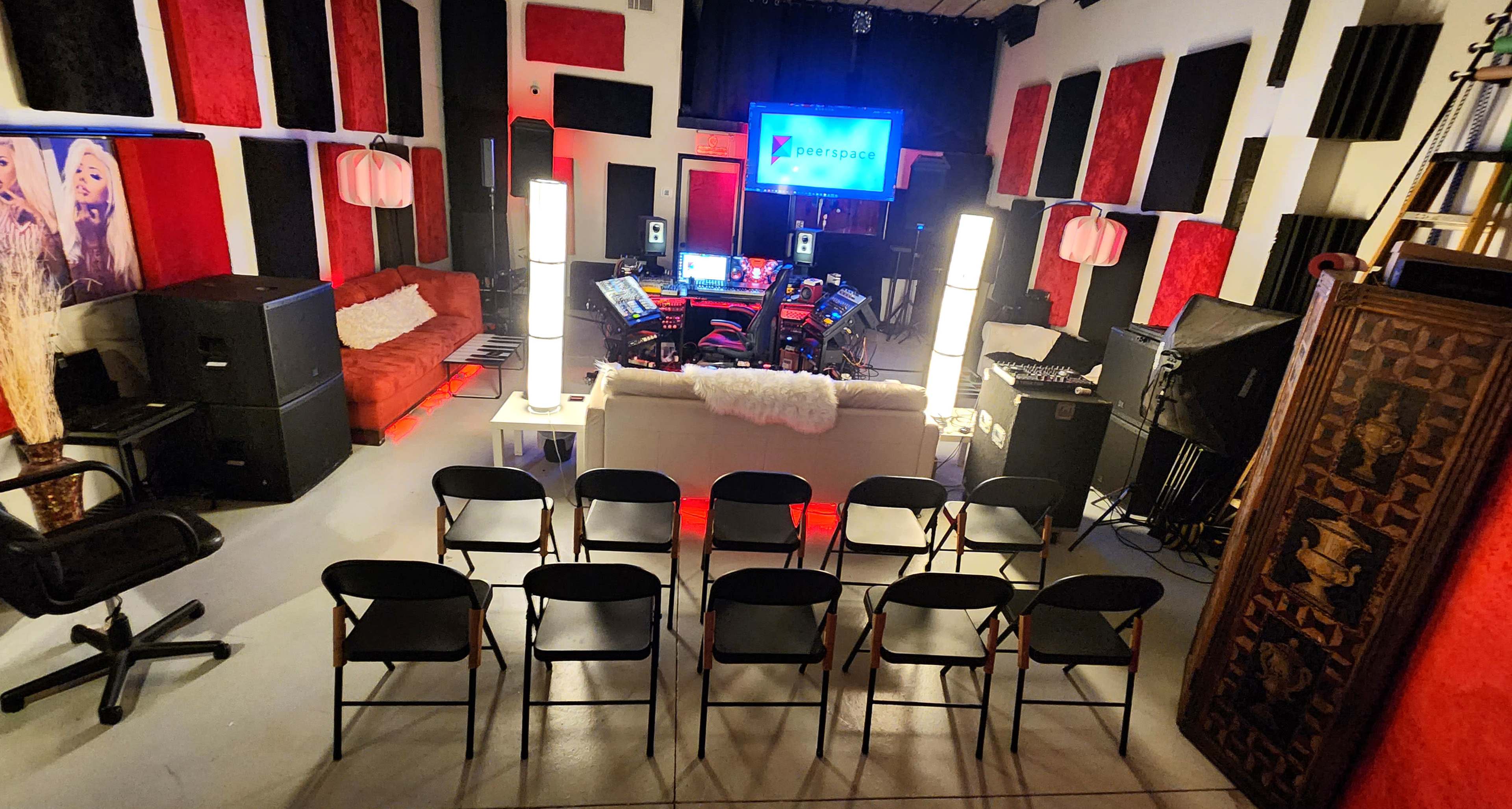 A music production studio features a central seating area with black chairs, equipment setups, and red and black soundproofing panels on the walls.