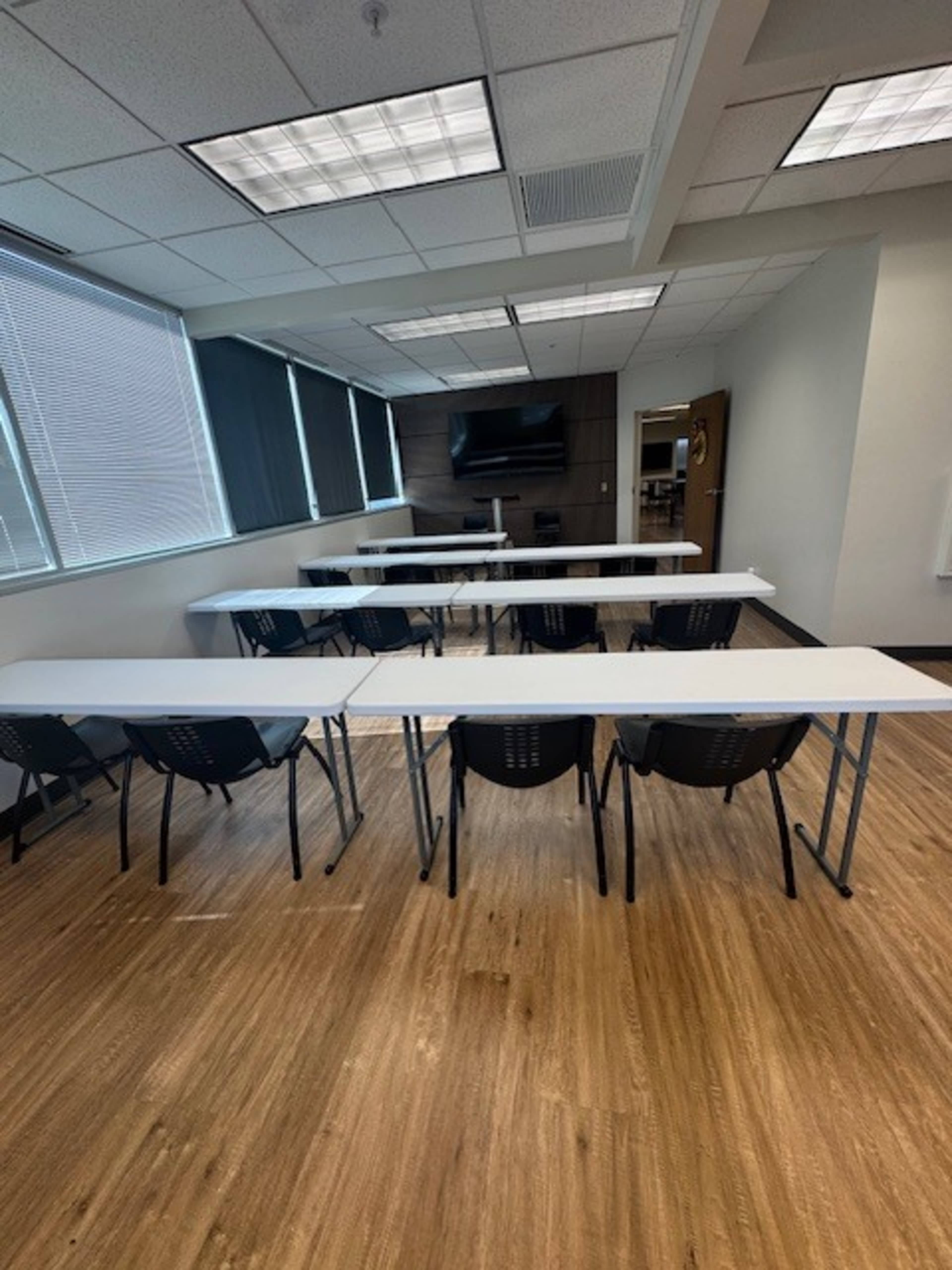 The image shows a conference room set up with multiple long tables arranged in a U-shape and chairs positioned around them, with a TV screen mounted on the wall.