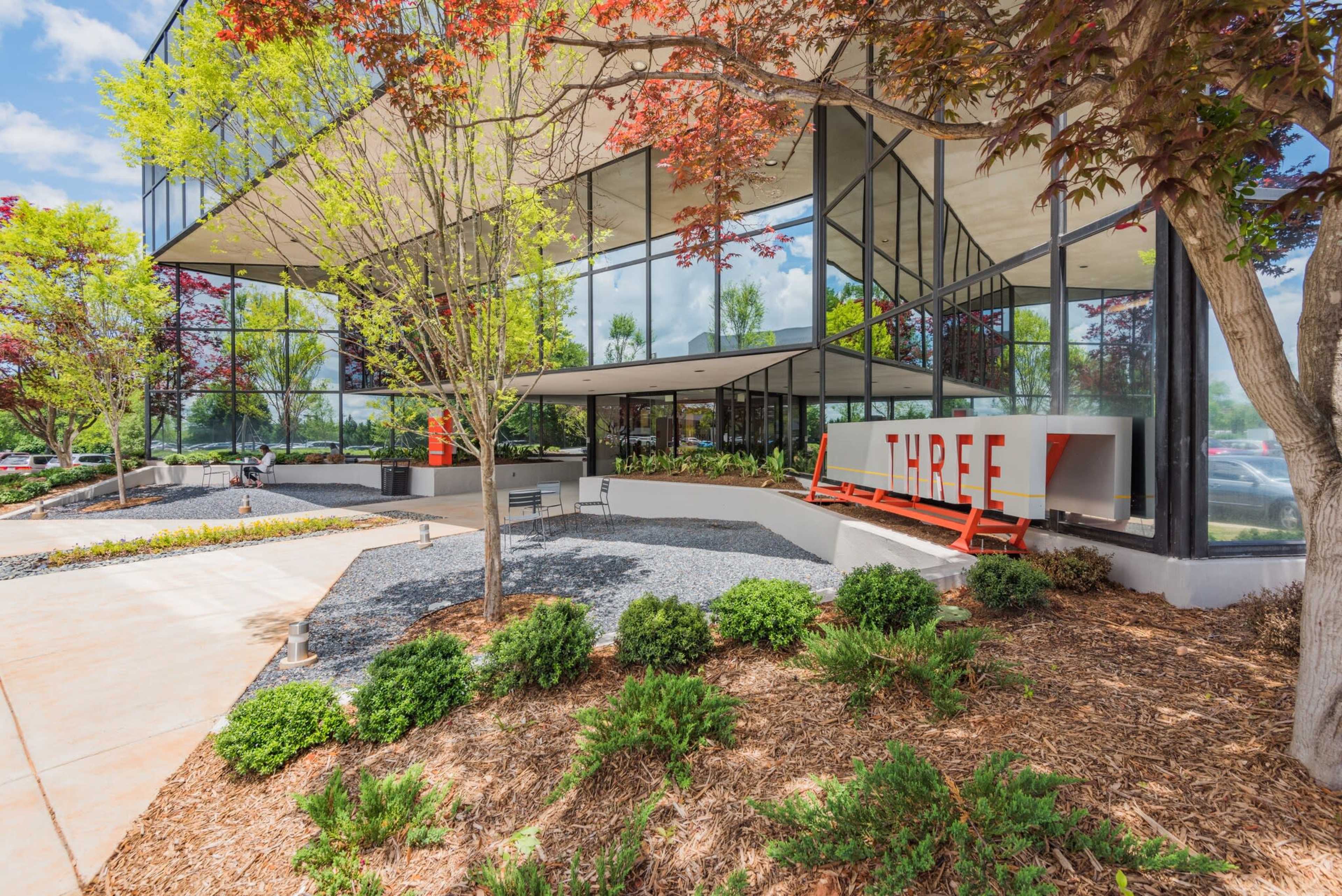 A modern office building features large glass panels, landscaped greenery, and a prominent sign reading "THREE" at the entrance.