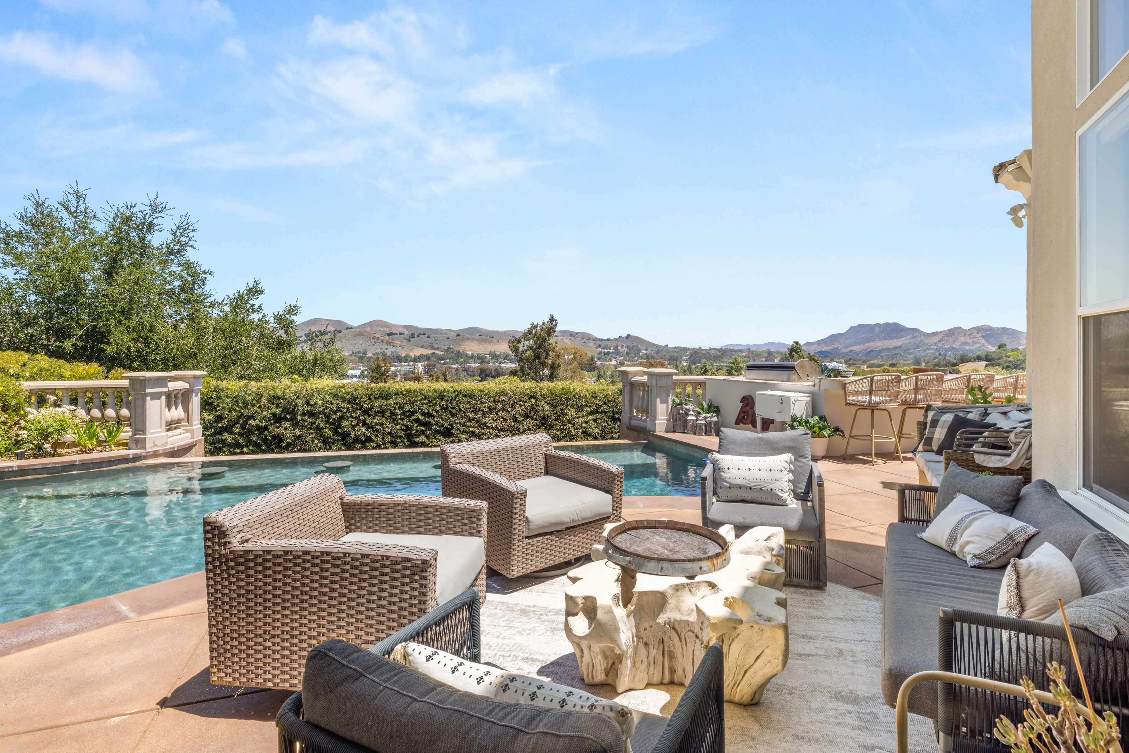 A patio area features a swimming pool surrounded by wicker seating and a fire pit, with a view of rolling hills and clear blue skies in the background.