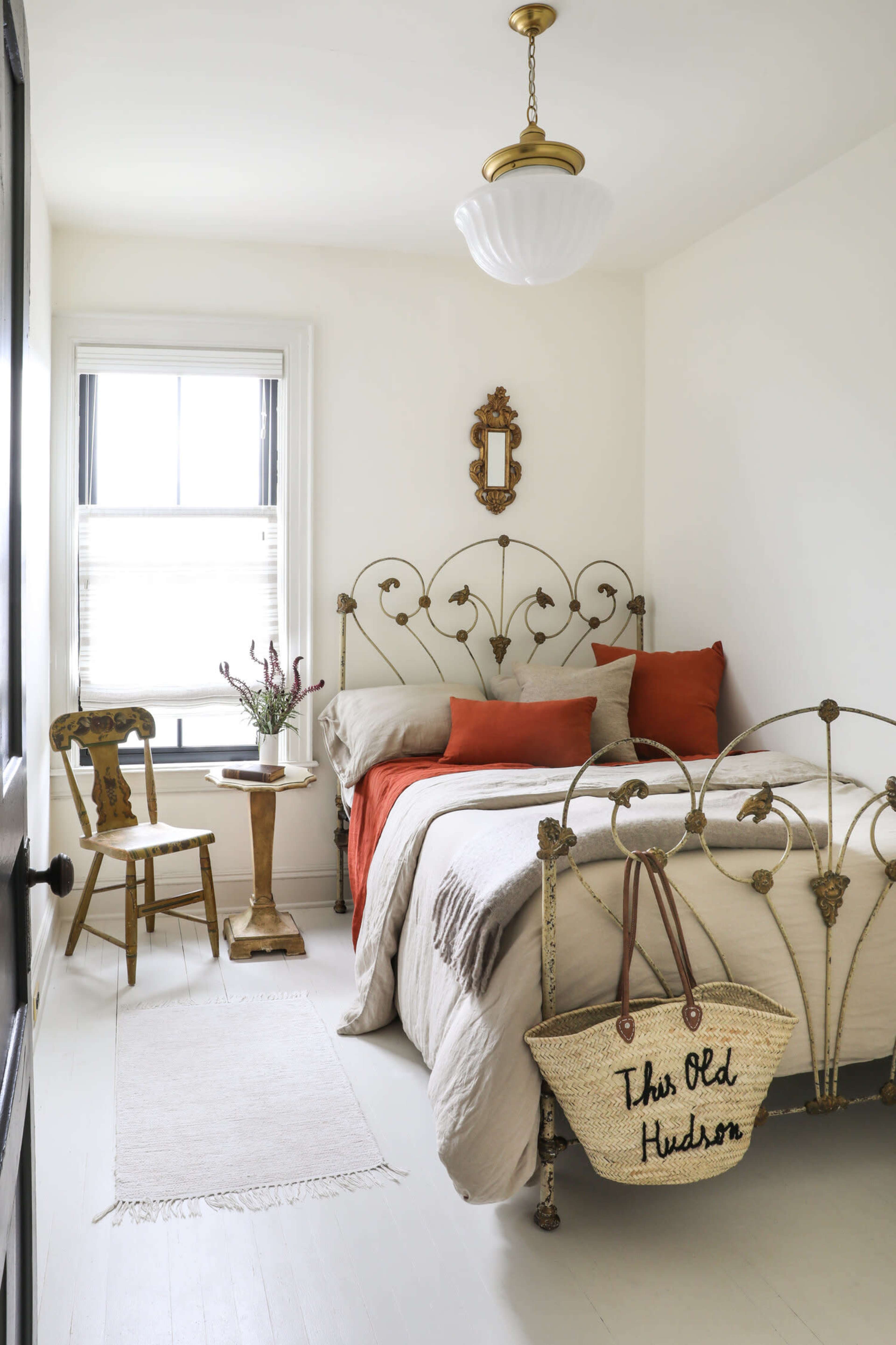 A small, light-colored bedroom featuring an ornate metal bed with orange pillows, a nearby wooden side table, a chair, and a woven bag resting against the bed.