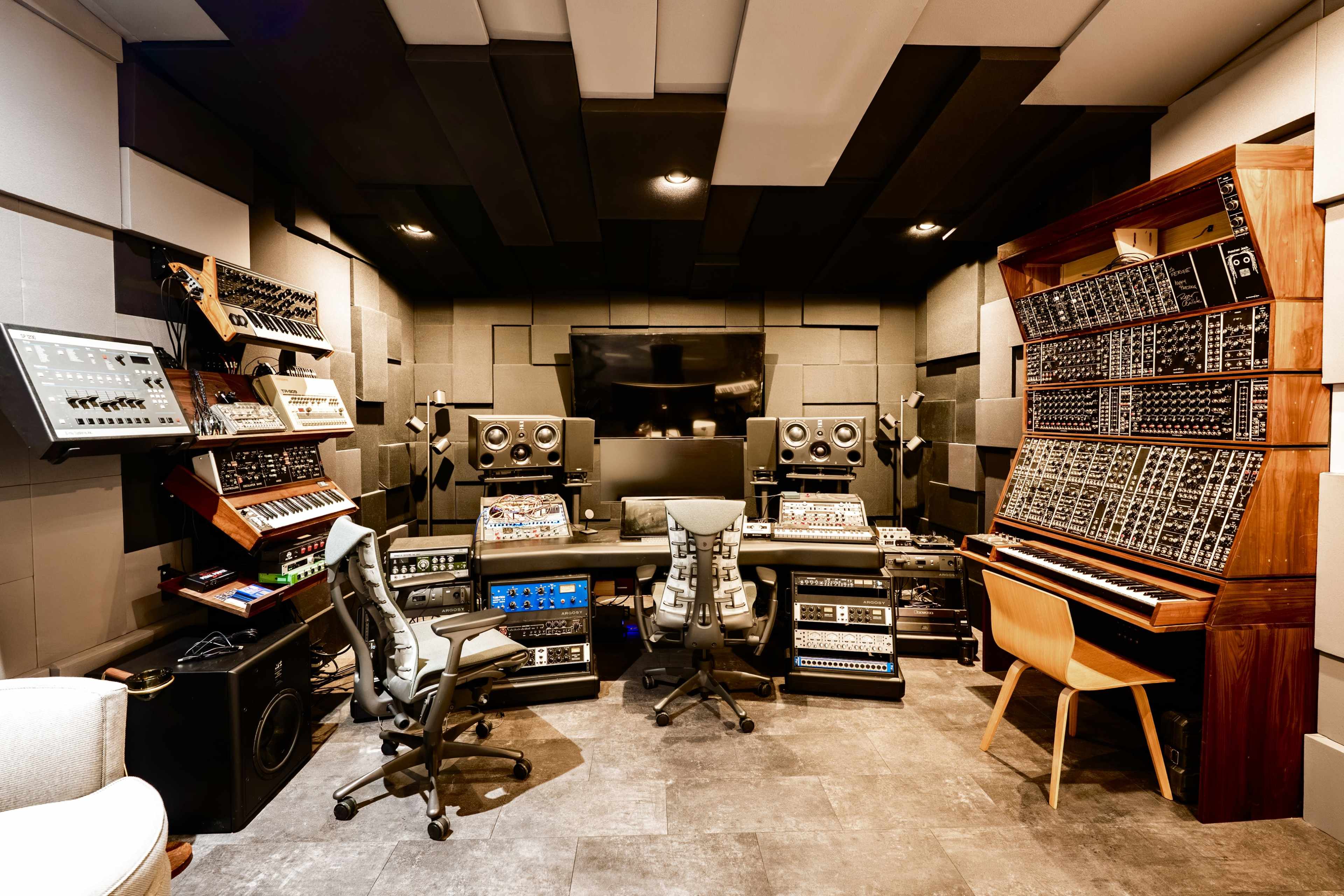 The image depicts a music production studio featuring various synthesizers, monitors, and mixing equipment arranged around a central workspace with modern furnishings.