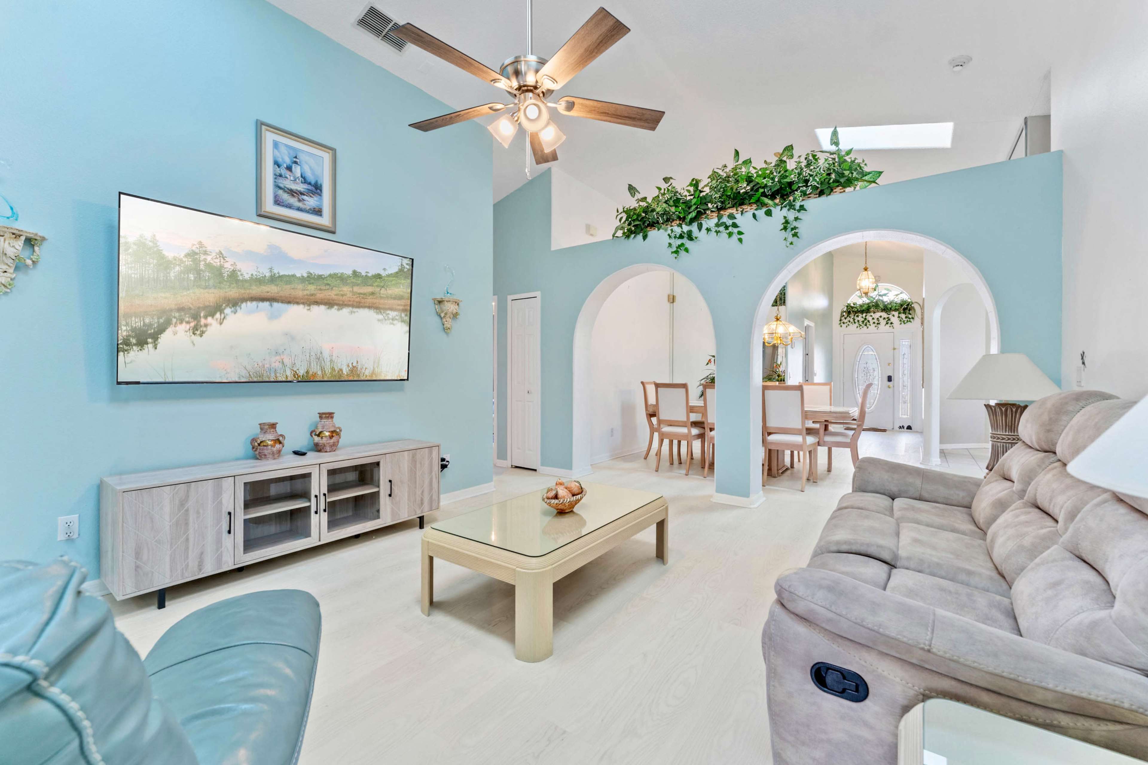 The image shows a bright living room with a sectional sofa, a television mounted on the wall, and an adjacent dining area featuring arches and greenery.