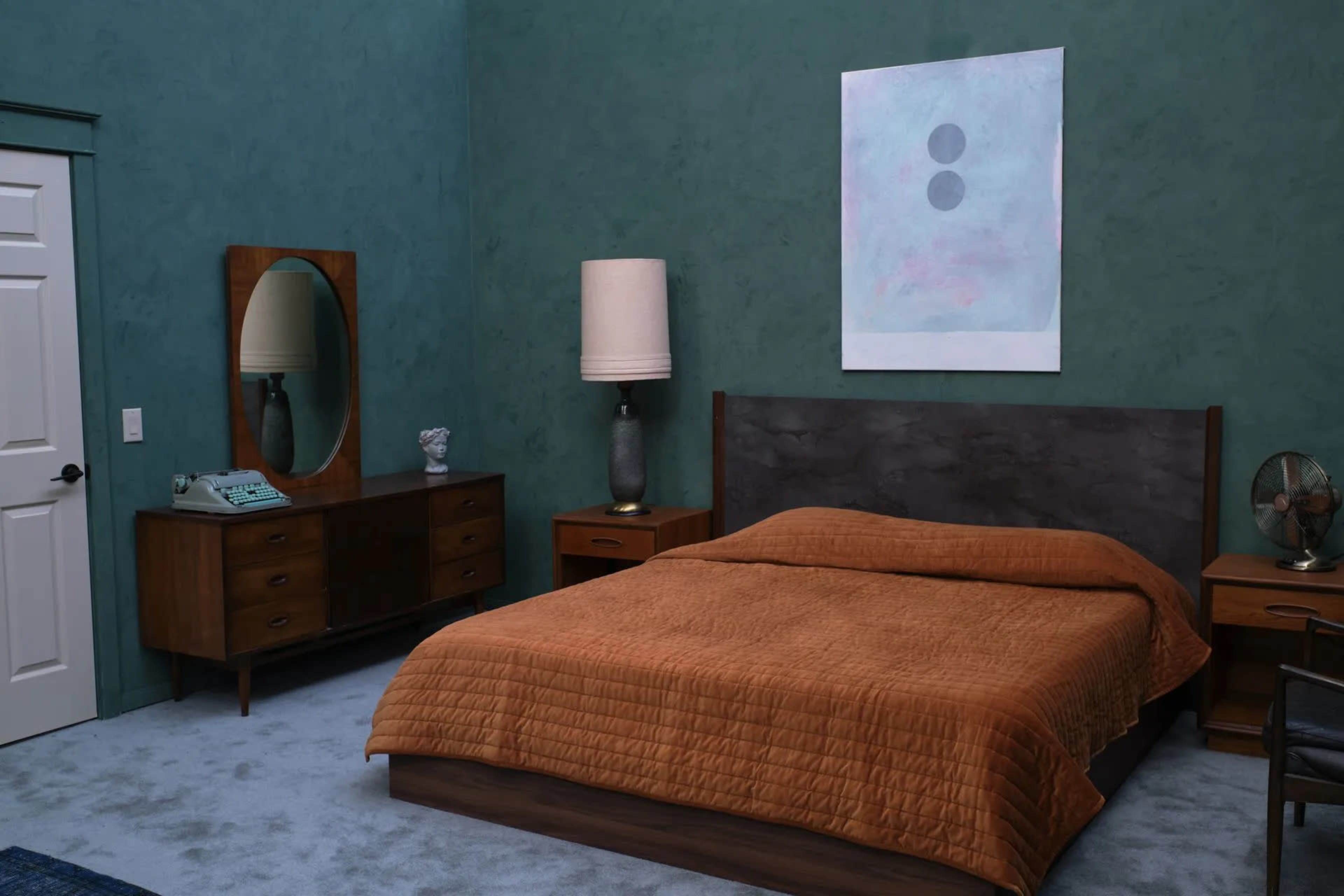 The bedroom features a large brown bedspread, a wooden dresser, a round mirror, and a modern abstract painting on the green wall.
