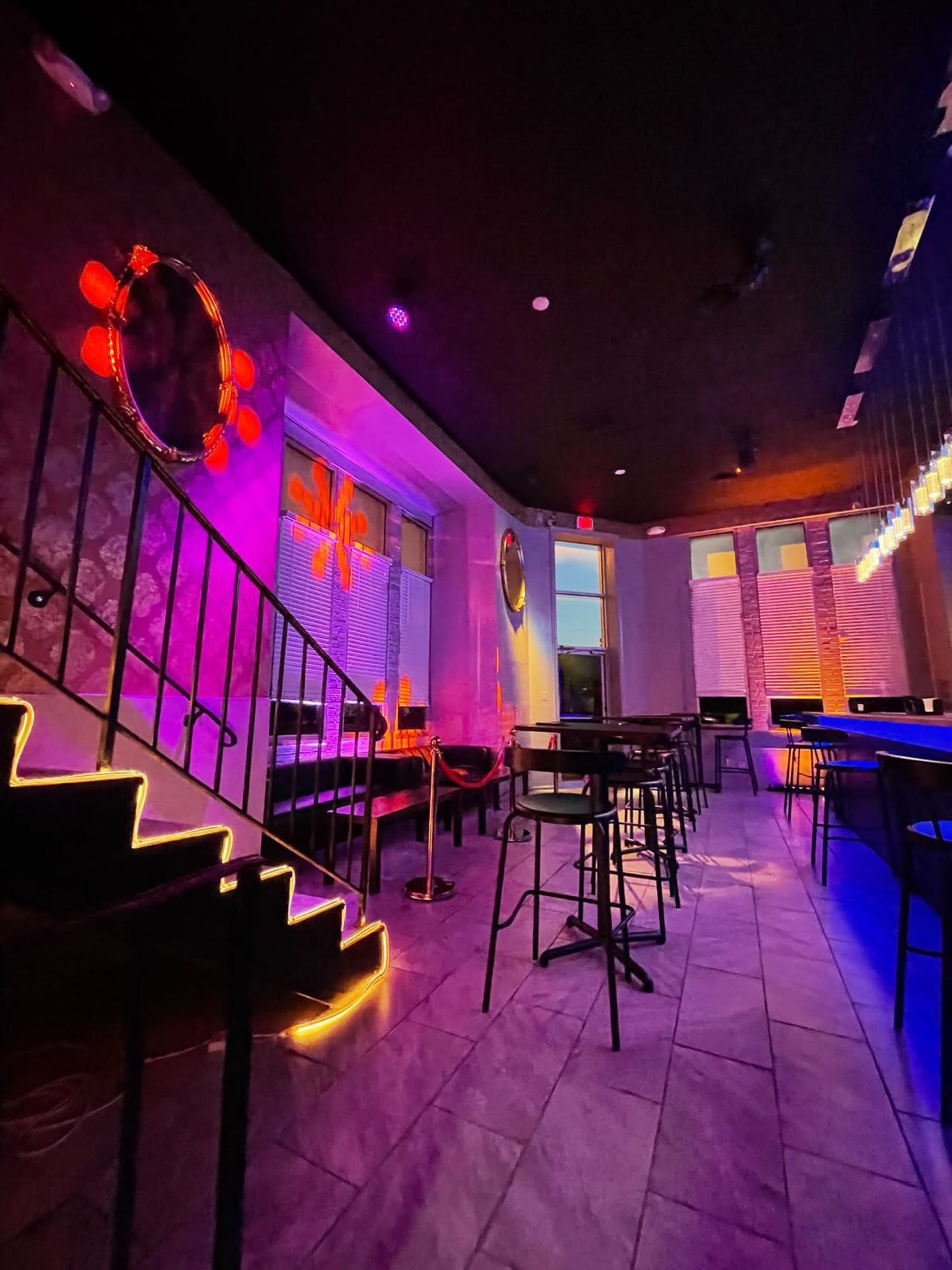 The image shows a dimly lit lounge with colorful lighting, a staircase leading up, and bar stools arranged near a countertop.