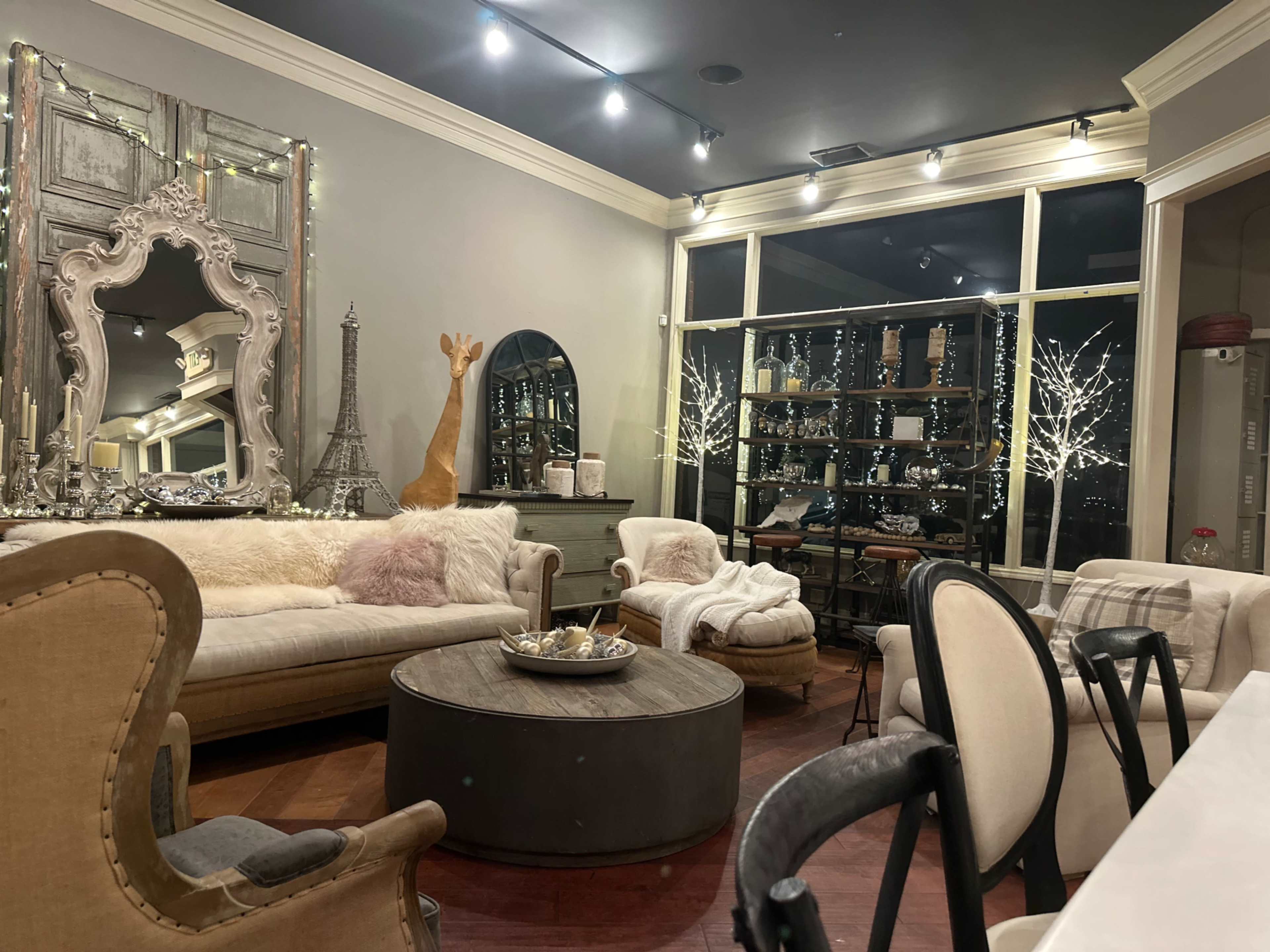 A cozy living space with a round coffee table, plush sofas, decorative mirrors, and a large window displaying twinkling lights outside.