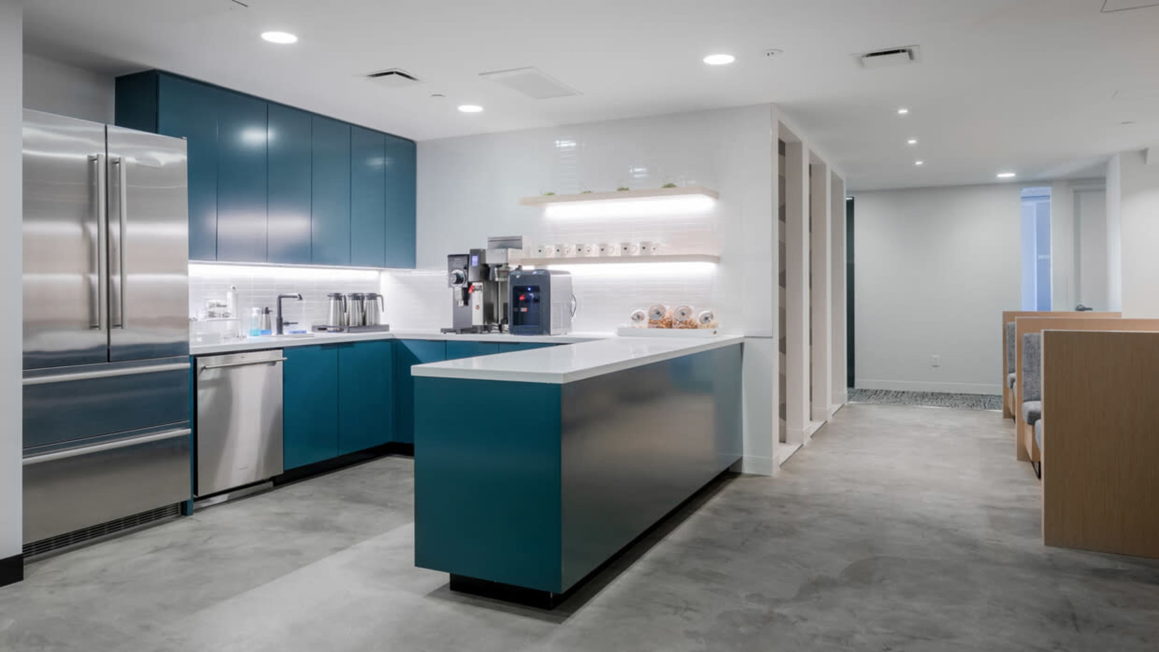 The image shows a modern kitchen featuring blue cabinetry, stainless steel appliances, and a white countertop with coffee machines and snack displays.