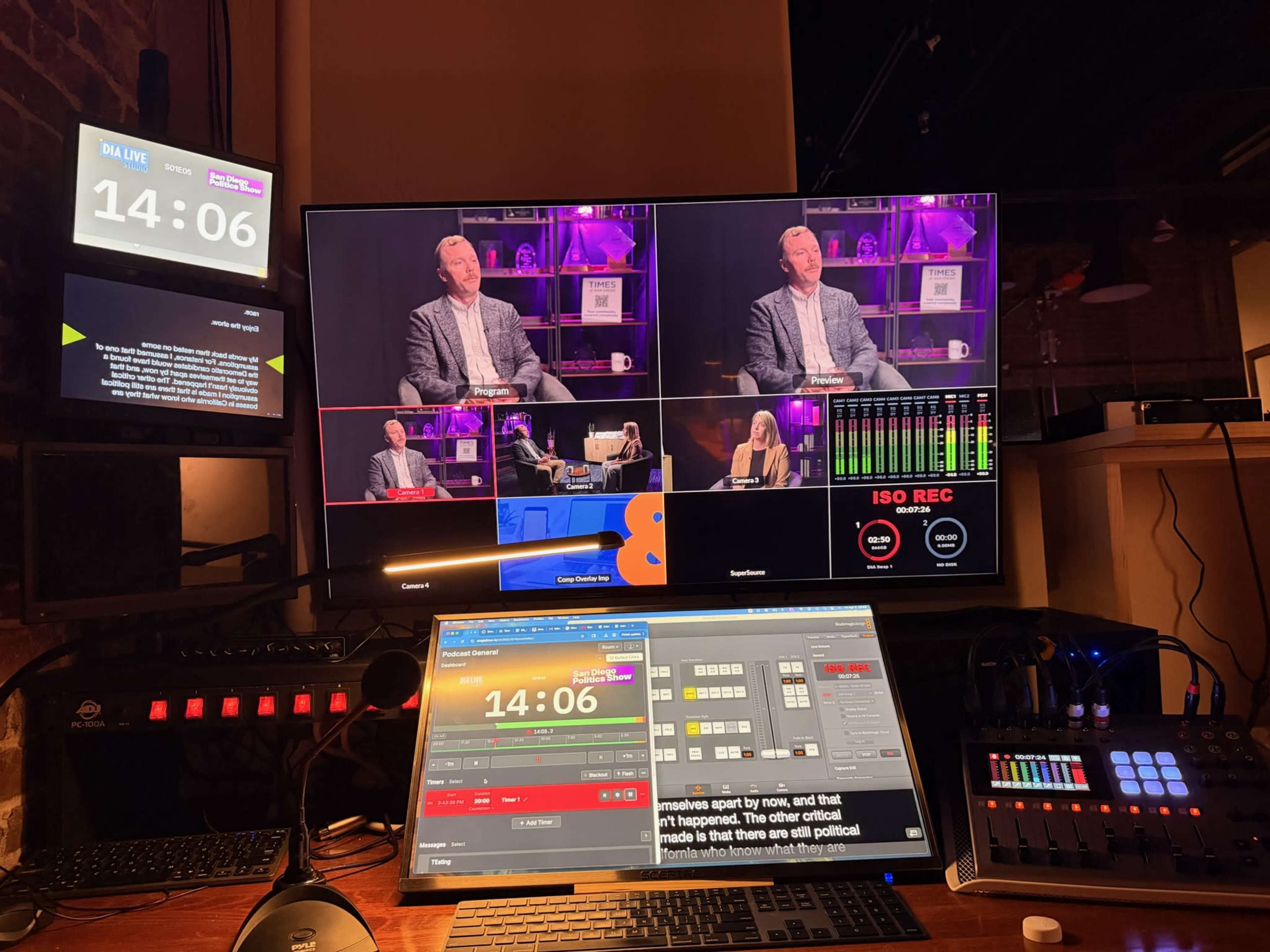 The image shows a television studio control room with multiple screens displaying a live interview, along with a control panel featuring various buttons and digital readouts.