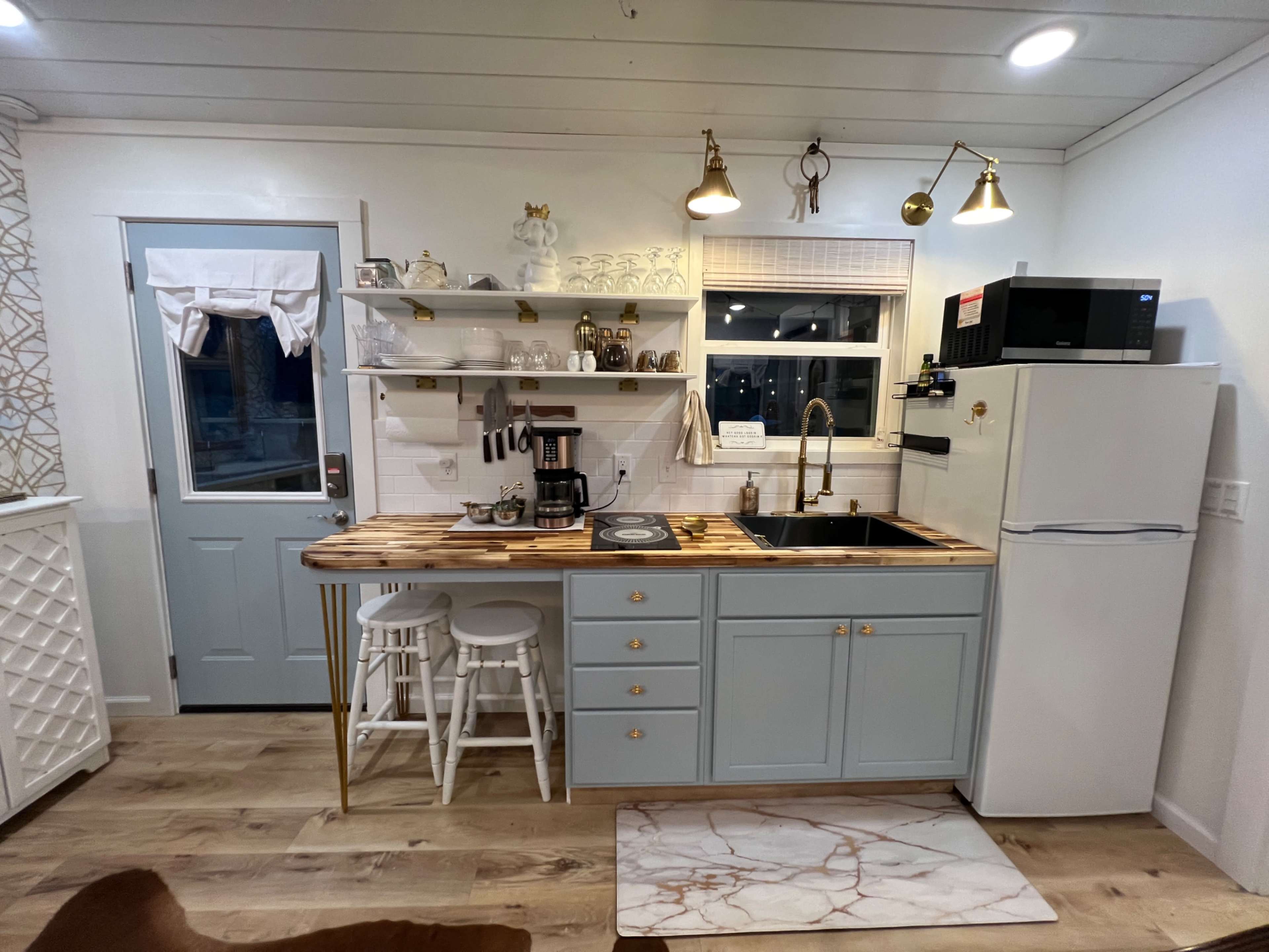 A small modern kitchen features light blue cabinetry, a white refrigerator, and open shelving with various kitchen items above a wood and black sink countertop.