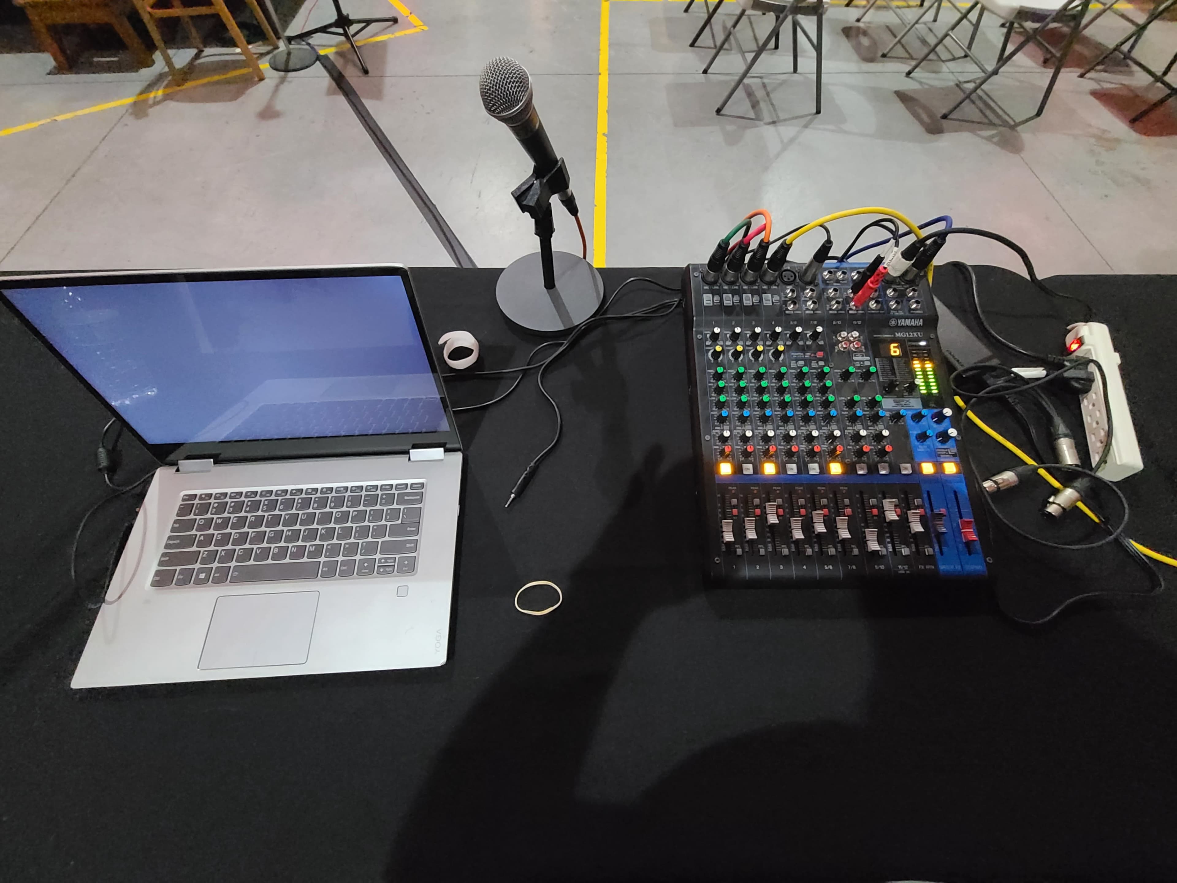 A laptop, a microphone, and an audio mixer are set up on a black table in a room with chairs arranged in the background.