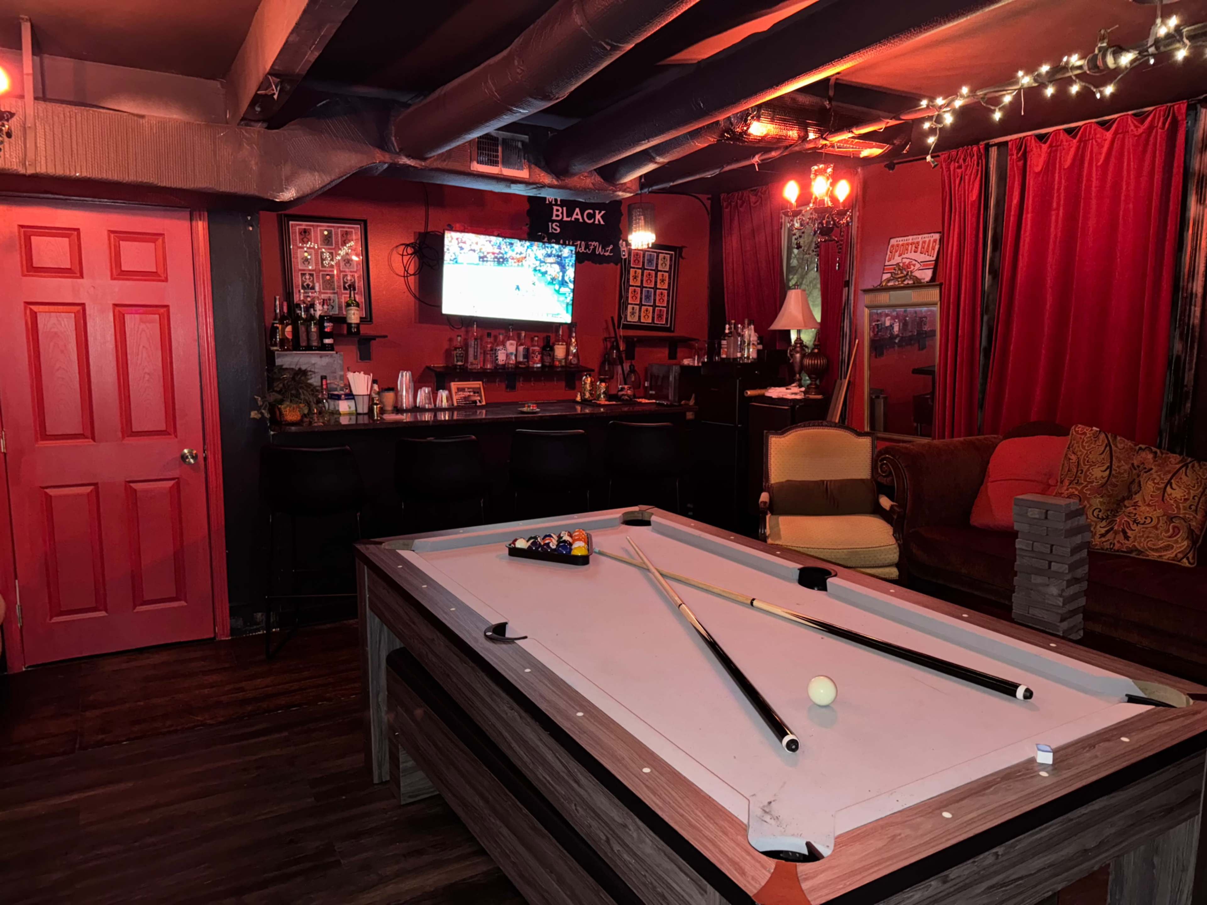 The image shows a dimly lit game room with a pool table in the center, a bar area in the background, and red walls decorated with various items and a television displaying a sports game.