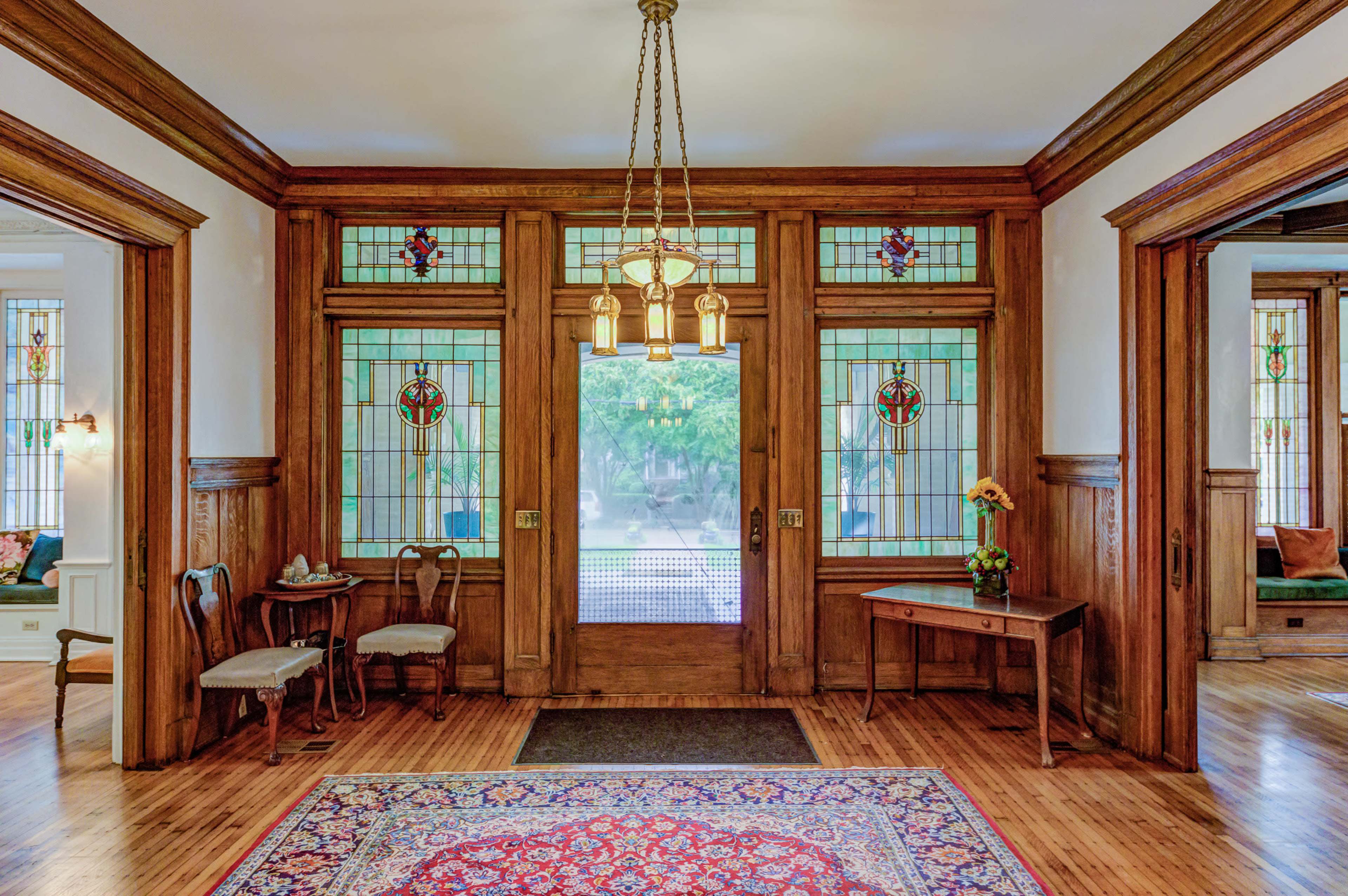 A spacious entryway features wooden walls, stained glass doors, a chandelier, and a decorative rug.