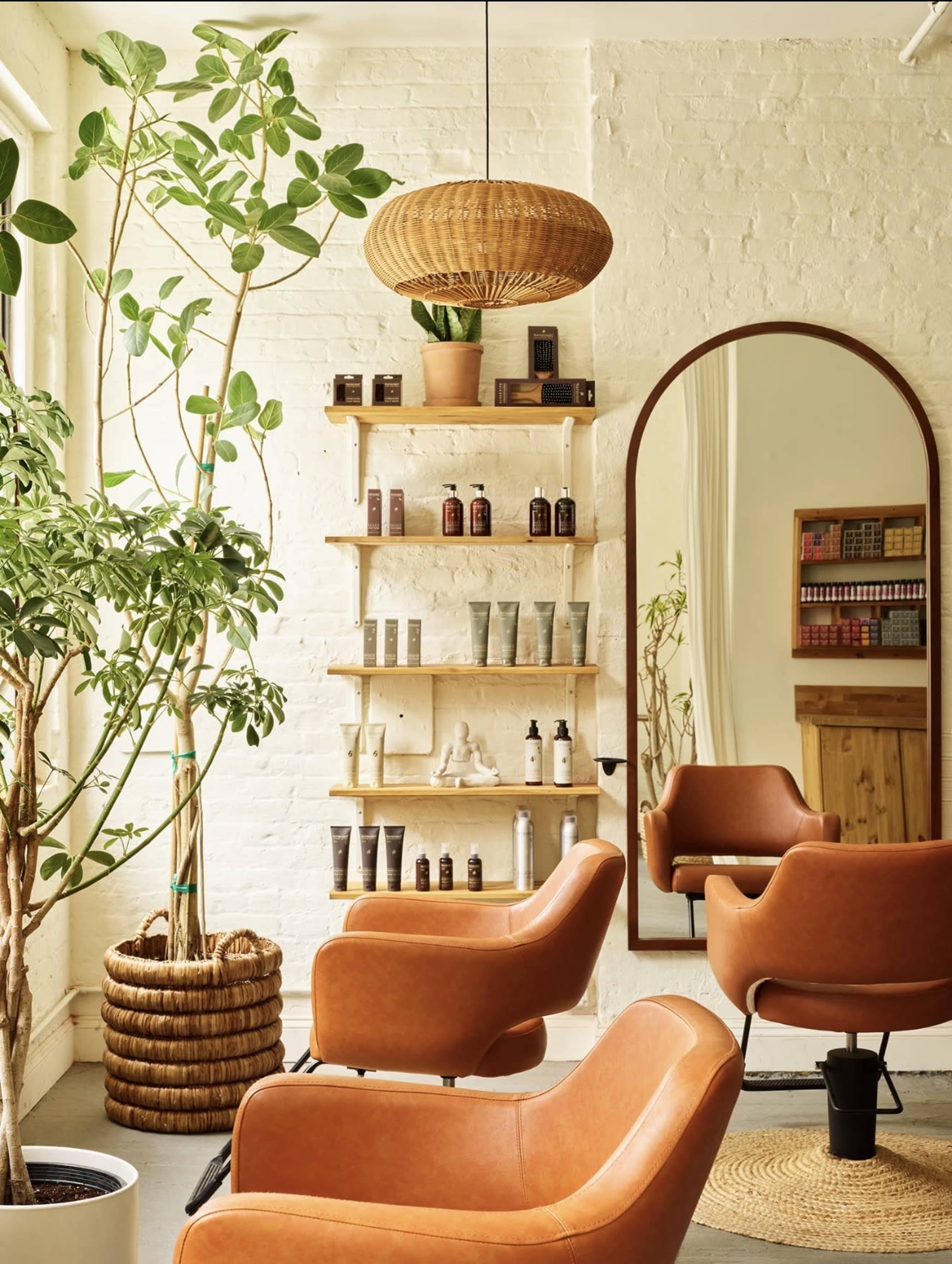 The image shows a modern hair salon featuring two orange chairs, a large mirror, a plant, and shelves displaying hair care products against a white brick wall.