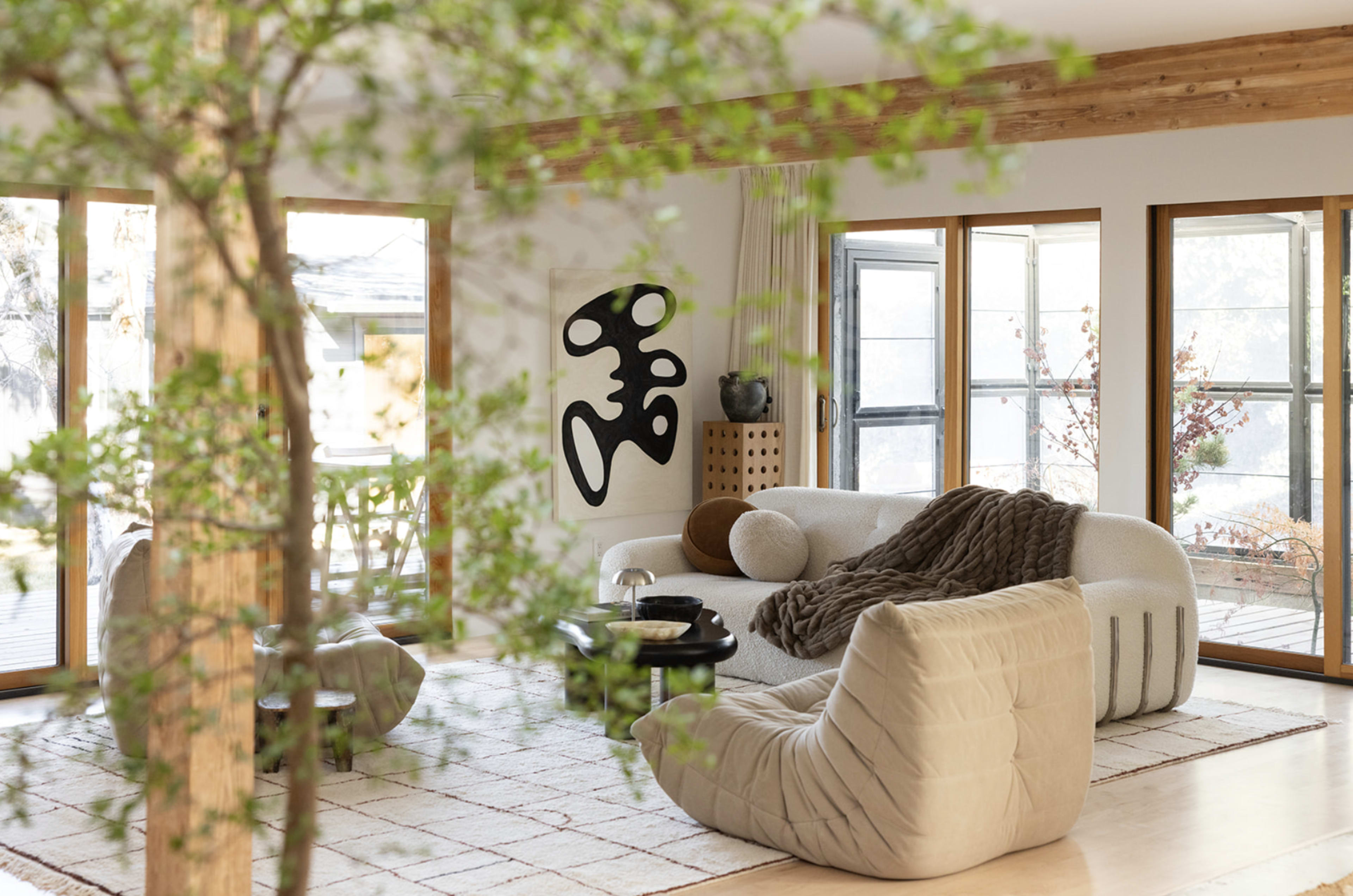 The image features a modern living room with a large, curved sofa, bean bag chairs, and a minimalist artwork on the wall, surrounded by large windows that let in natural light.