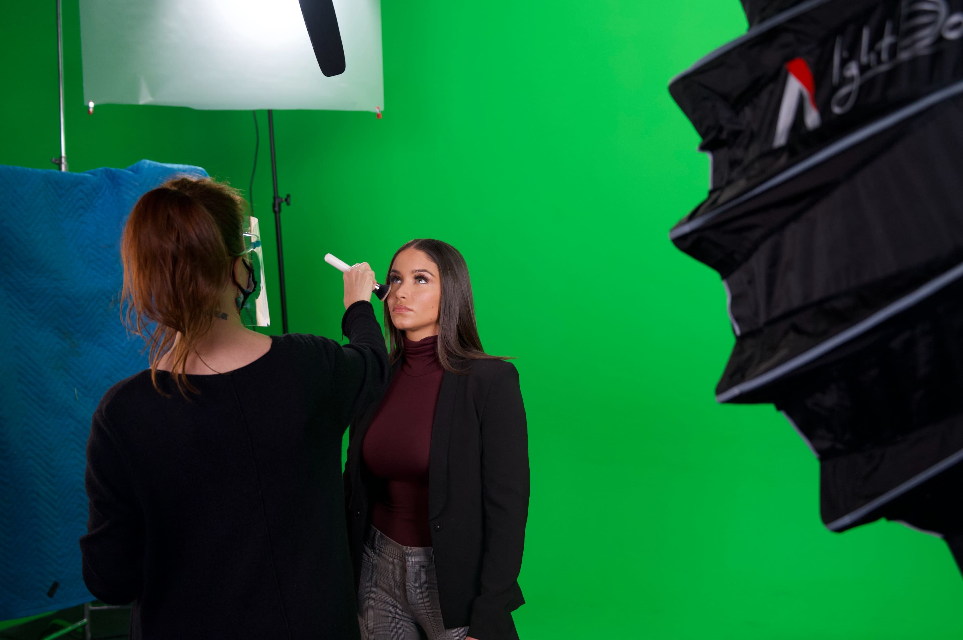 A woman is being prepared for a photo shoot in front of a green screen, while a stylist adjusts her makeup.