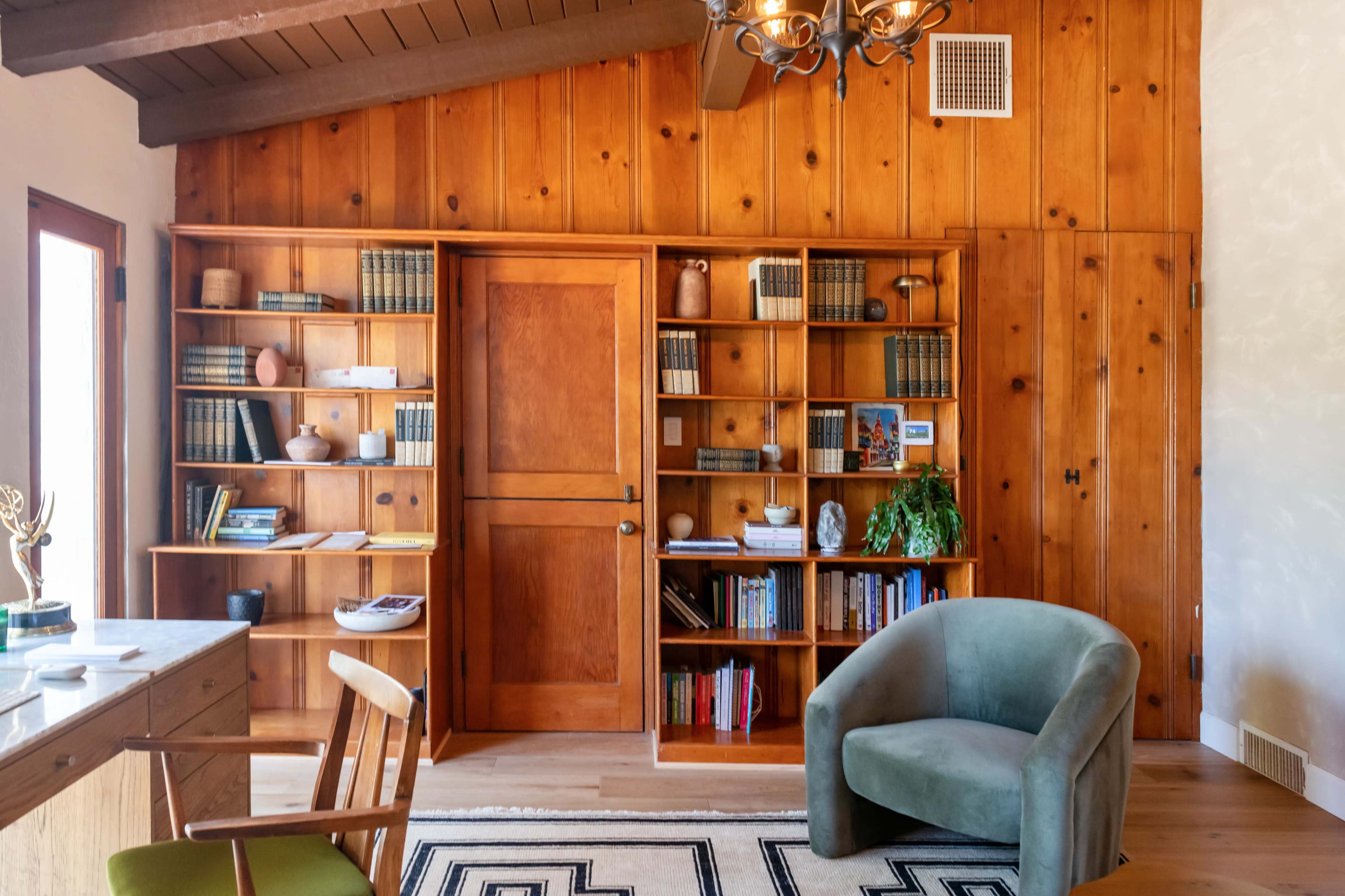 The image shows a room with wooden paneled walls, a bookshelf filled with books and decorative items, and a green upholstered chair next to a wooden desk.