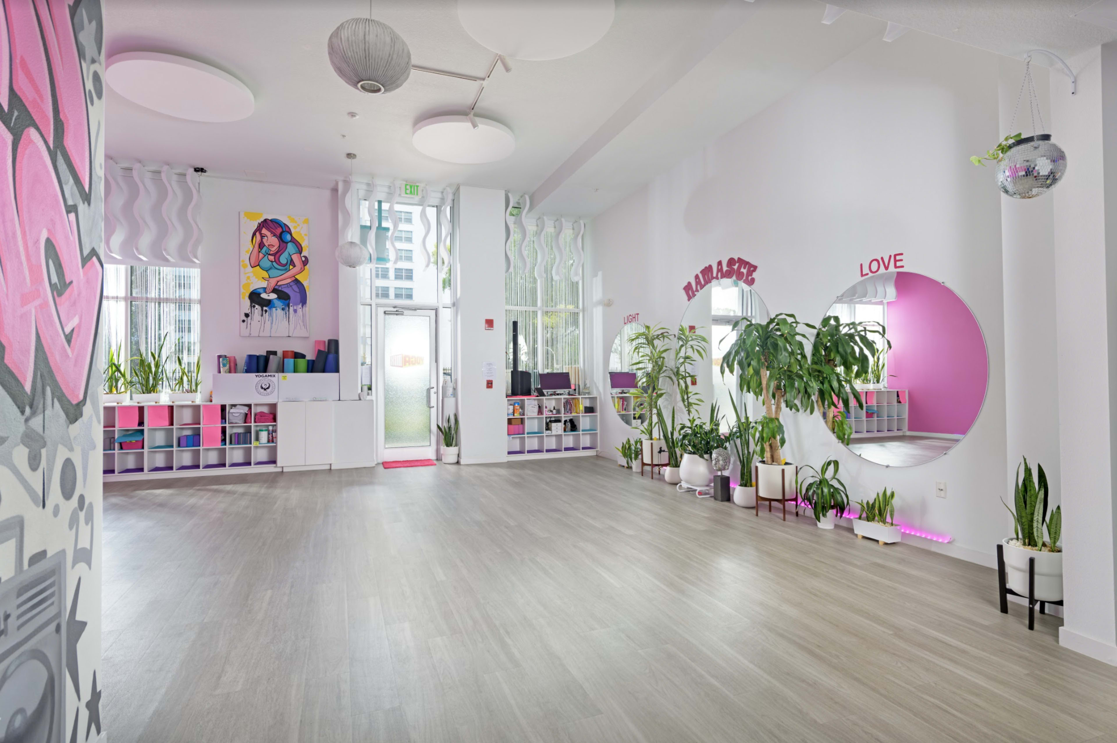 A spacious, well-lit room features large windows, colorful wall art, and indoor plants, with areas designated for storage and mirrors.