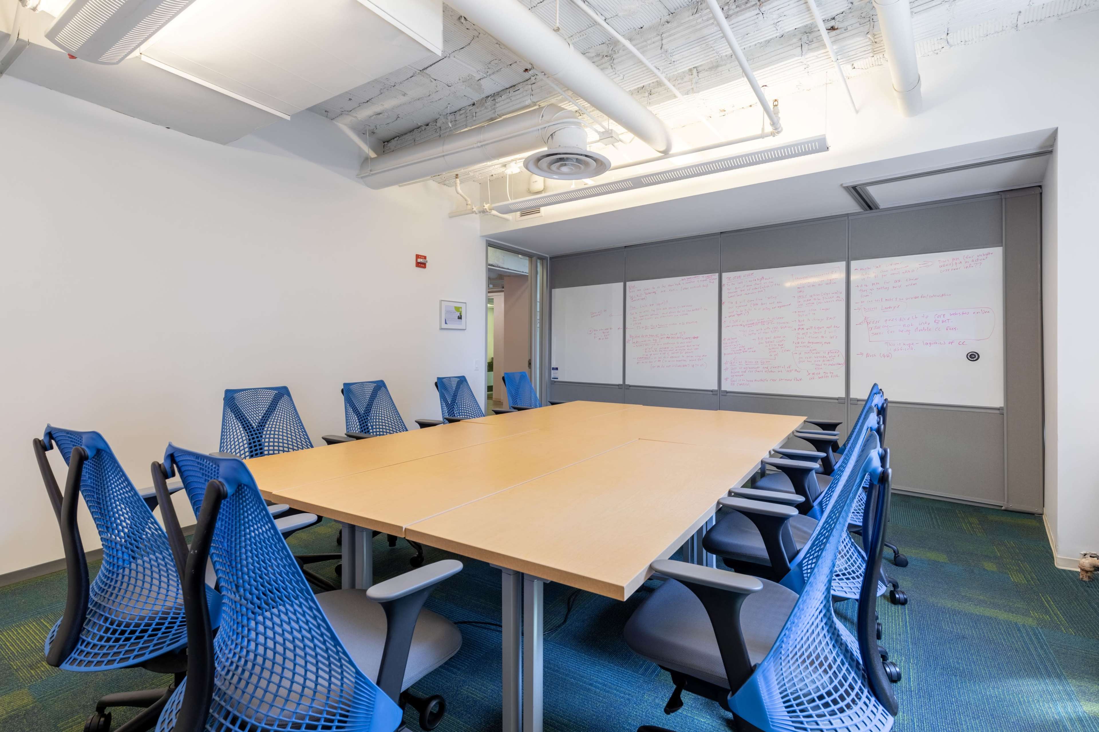 A conference room features a large rectangular table surrounded by blue chairs and a wall with whiteboards covered in notes.