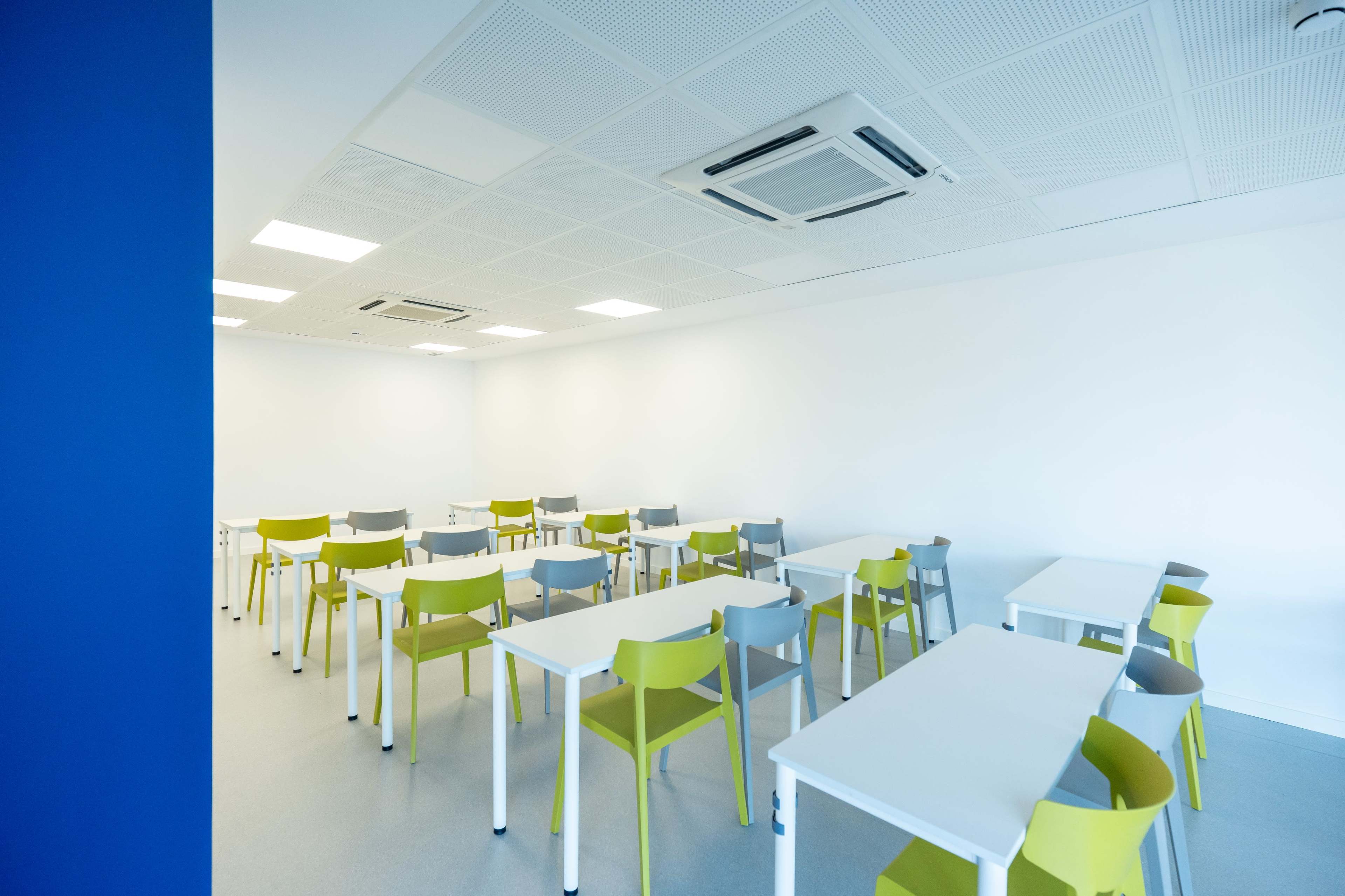 The image shows a modern classroom with white walls, a light gray floor, and rows of white tables surrounded by colorful chairs in shades of green and gray.