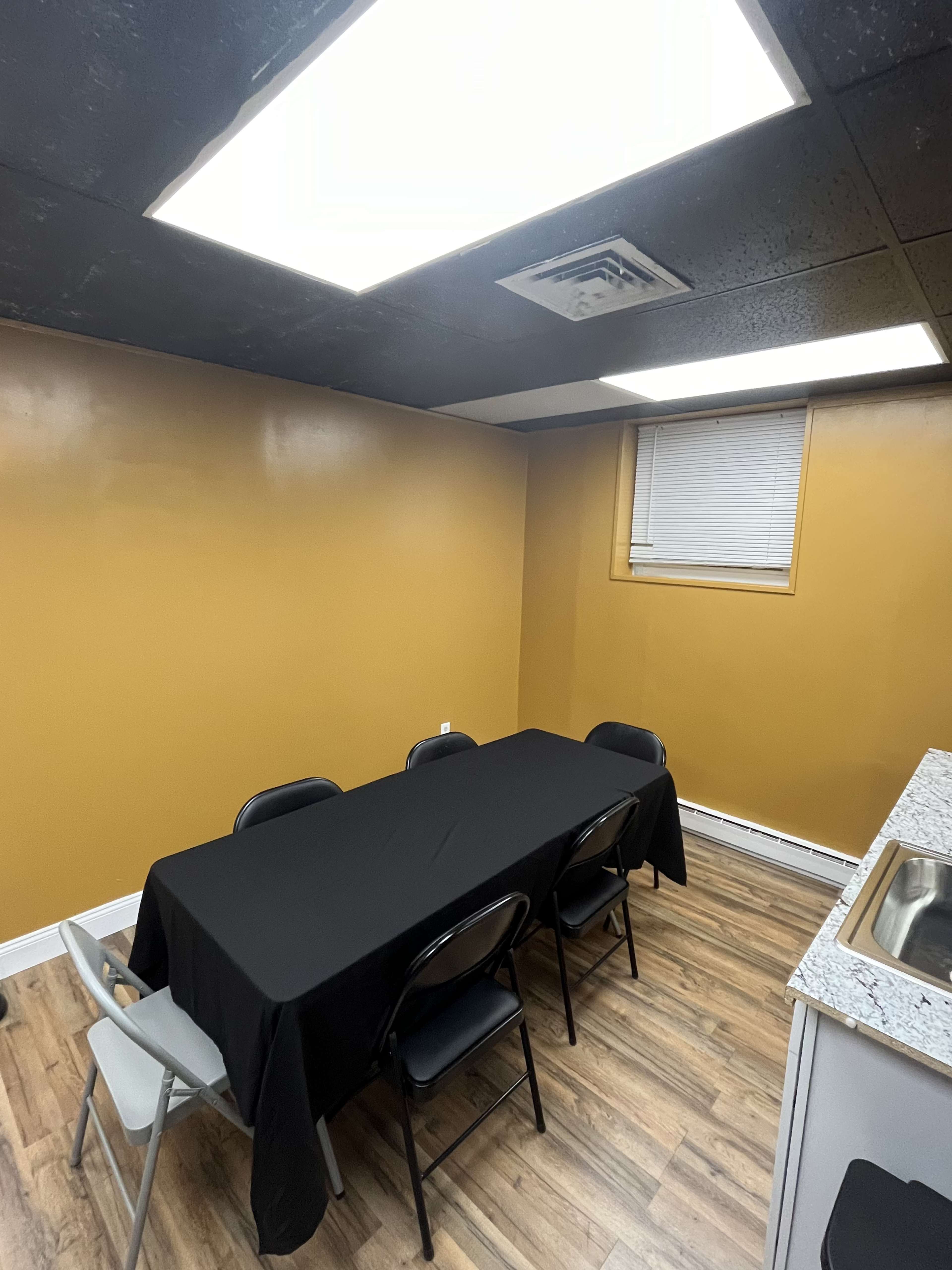 The room features a rectangular table covered with a black cloth surrounded by six chairs, with yellow walls and fluorescent ceiling lights.